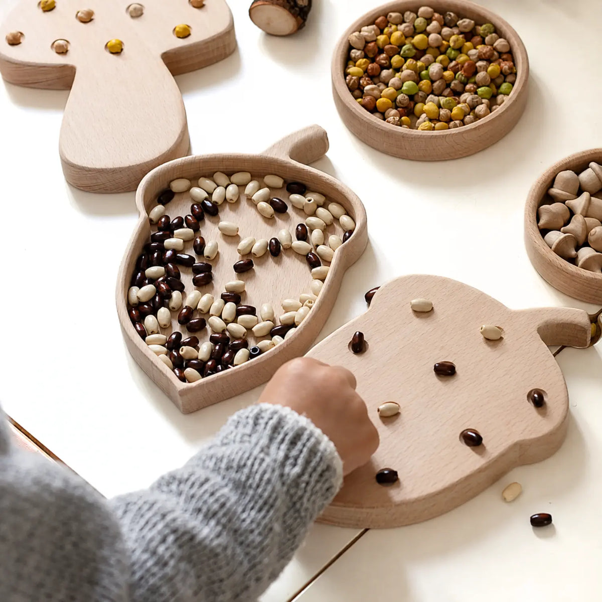Acorn-shaped wooden sorting tray with black and white beans beside a child sorting natural loose parts on a tabletop.
