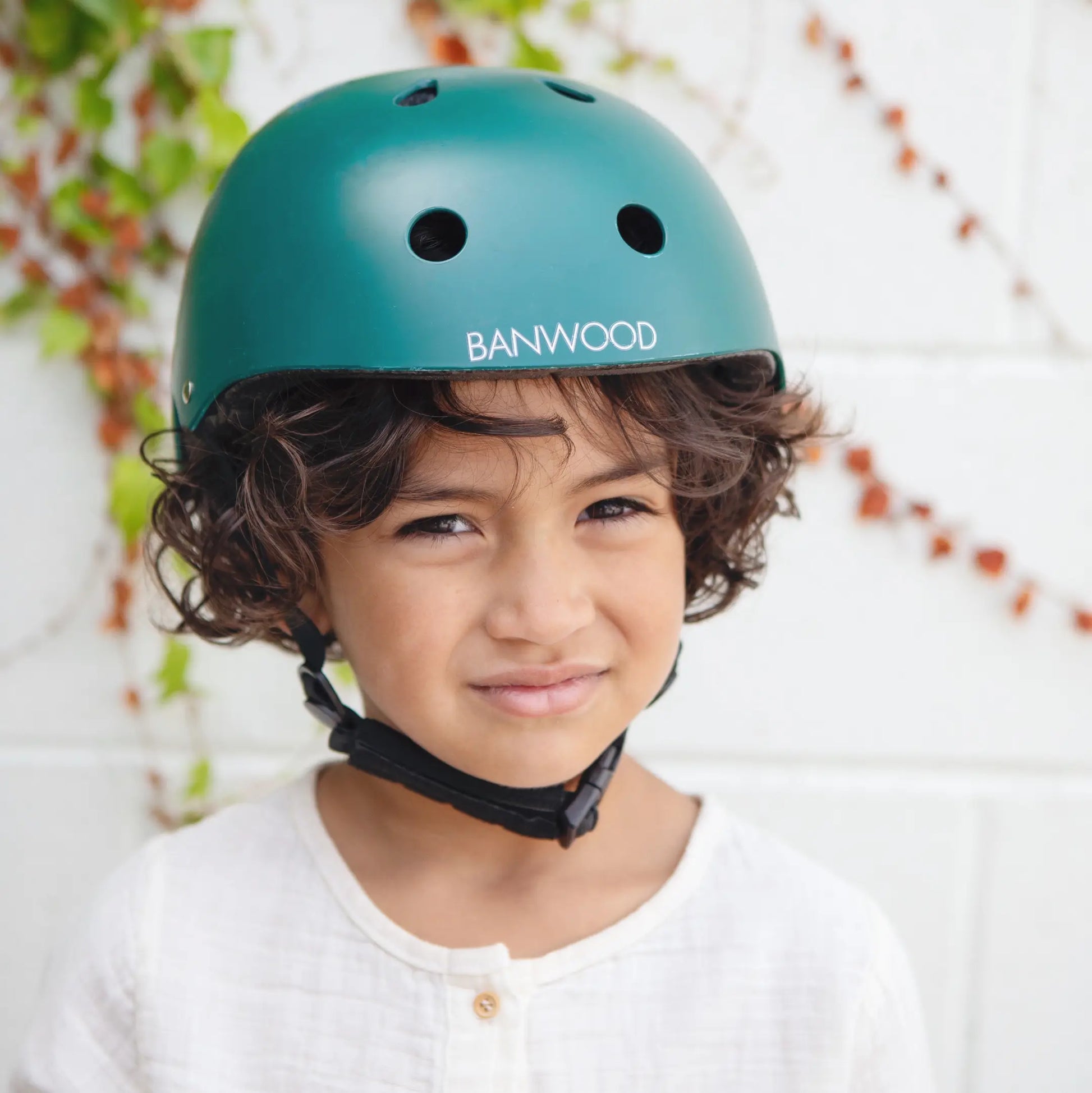 Child wearing a  dark green bicycle helmet with Banwood branding against a blurred natural background