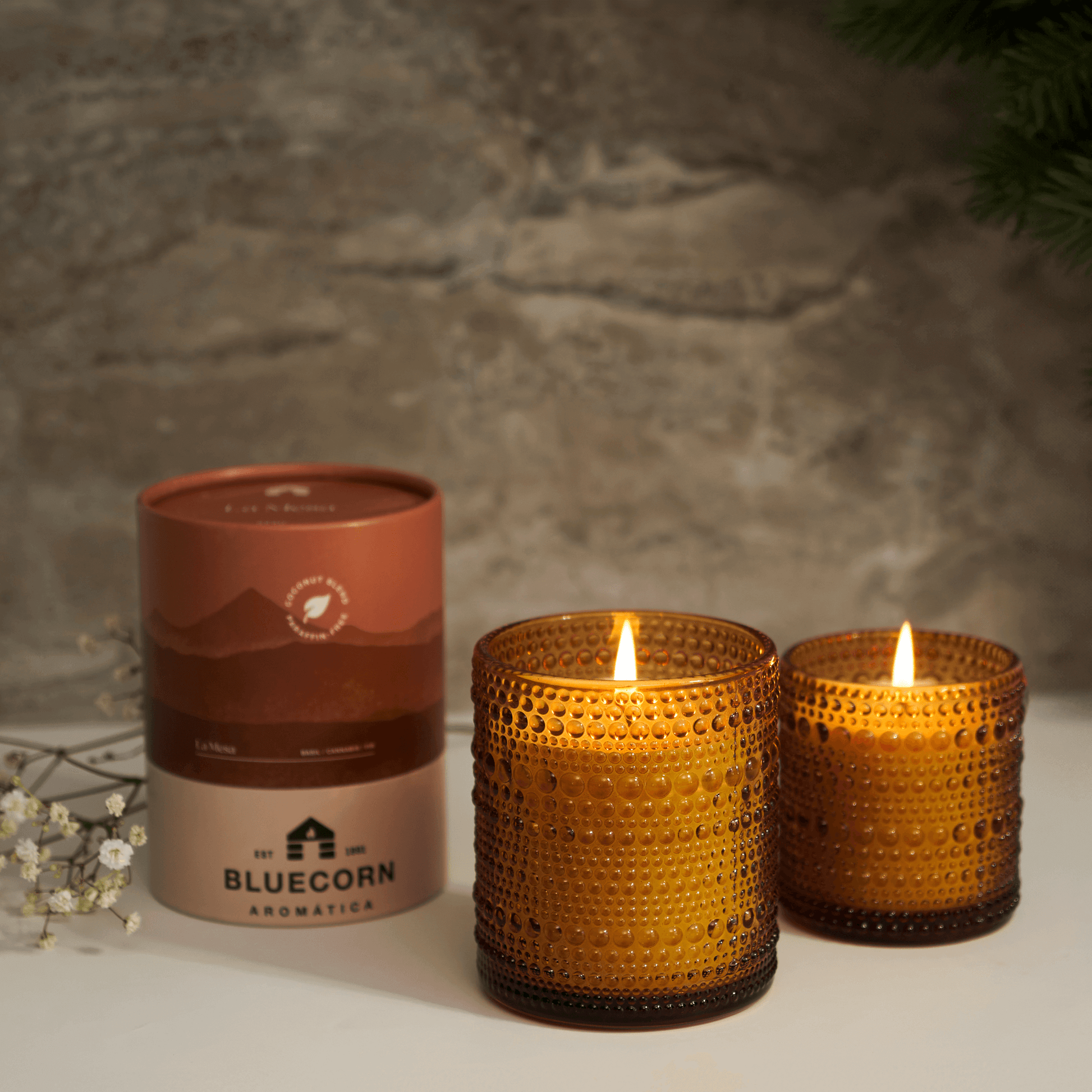 Two lit amber-colored candles with a cylindrical packaging labeled 'Bluecorn' on a neutral background.