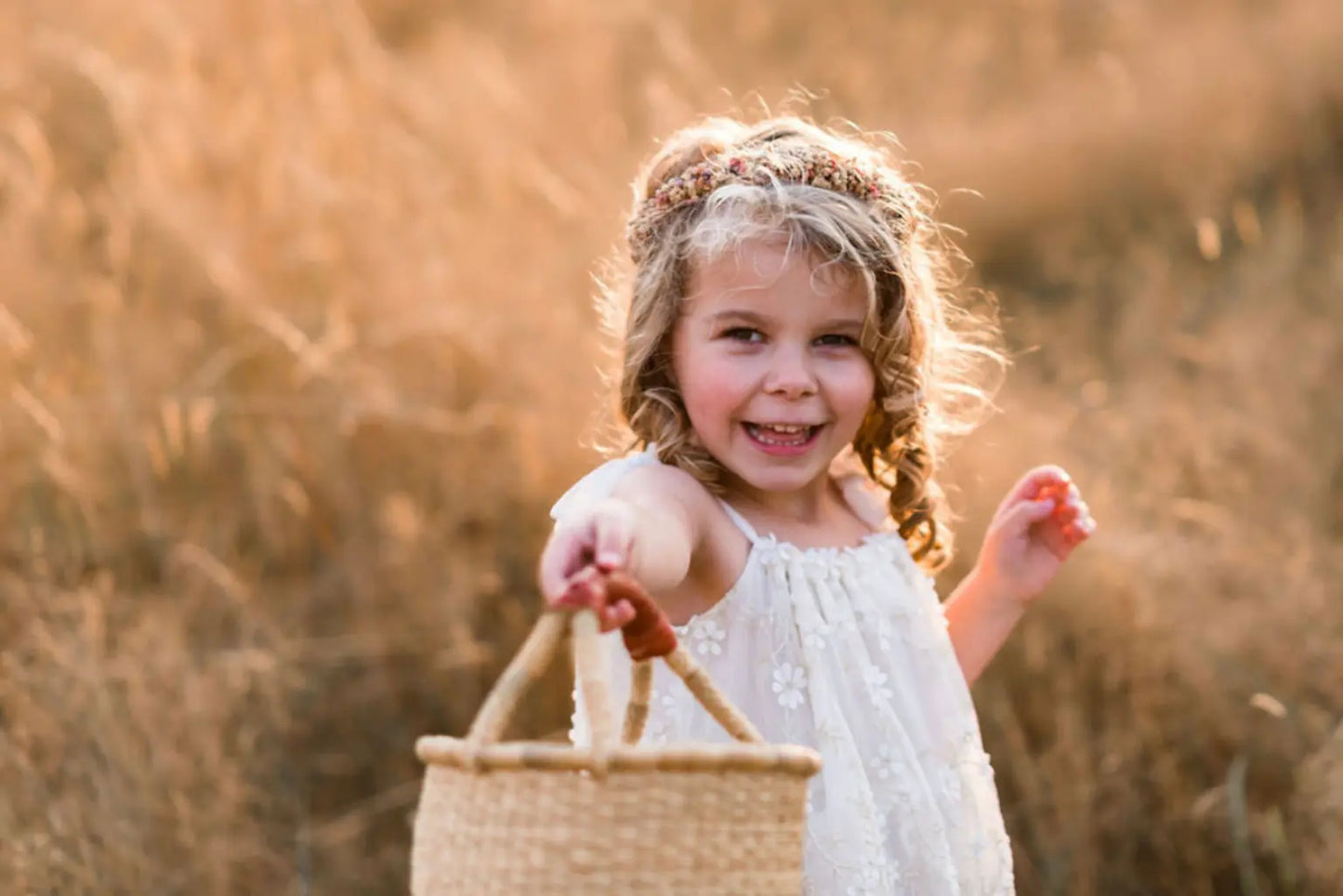Young girl in a white dress holding a woven basket in a field of tall grass.