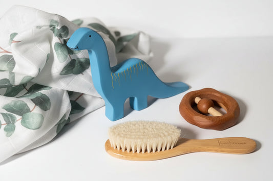 Blue dinosaur toy, wooden brush, and wooden ring on a wooden surface with white and green baby swaddle.