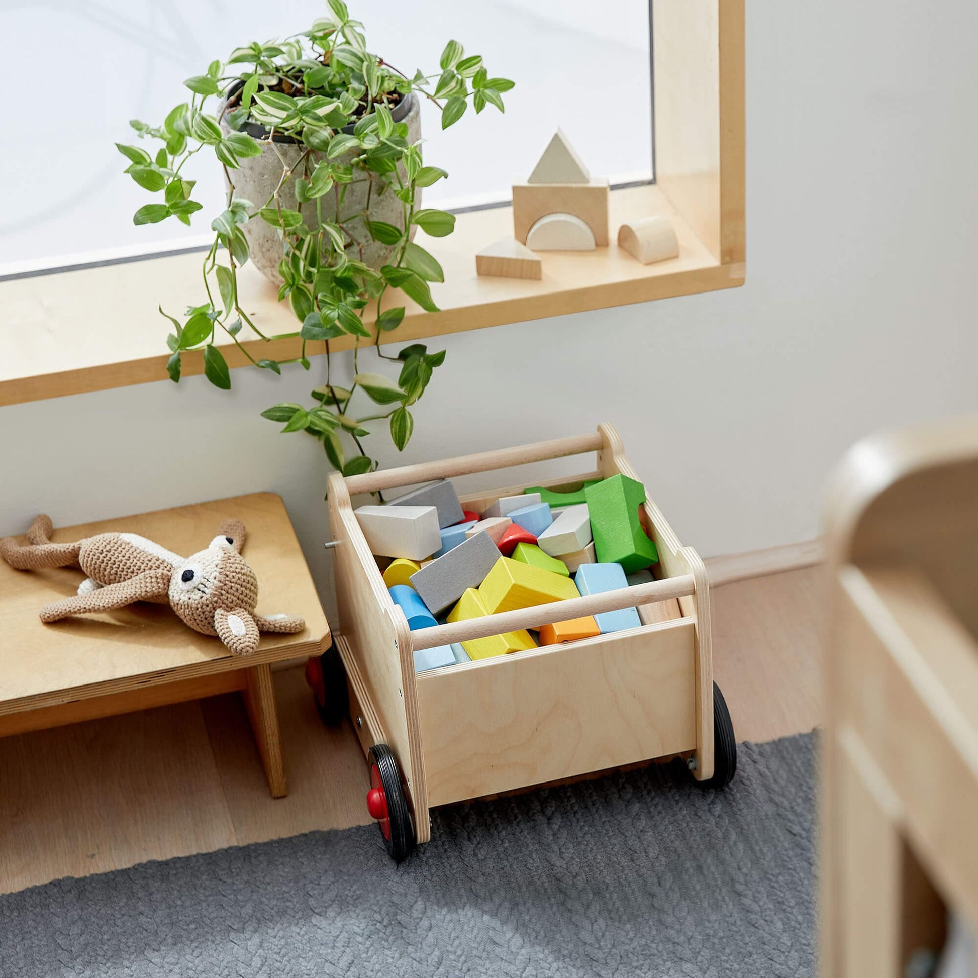 Wooden toy cart filled with colorful blocks in a room with a shelf and plant.
