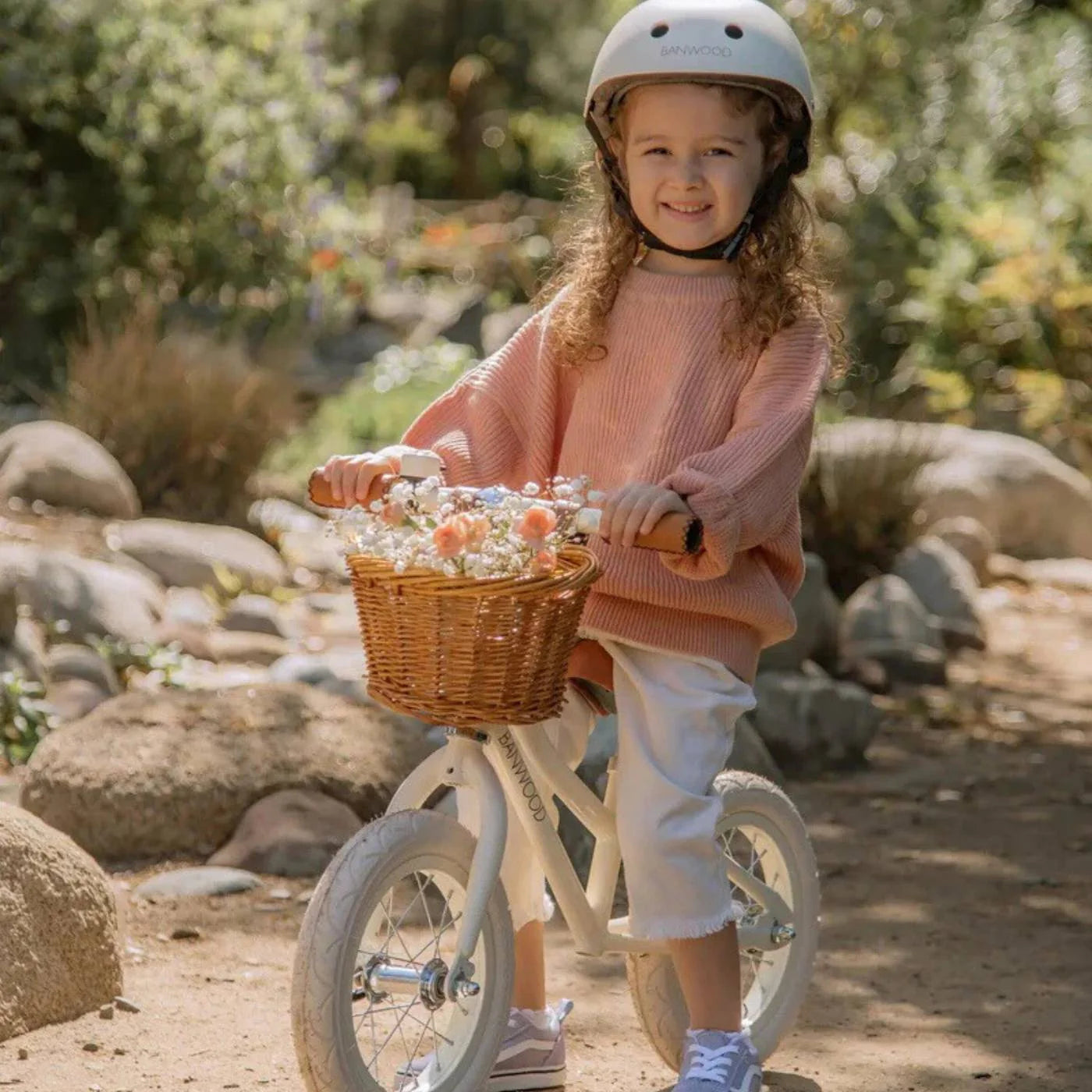 Child on a bicycle with a basket full of flowers, wearing a helmet in a natural setting.