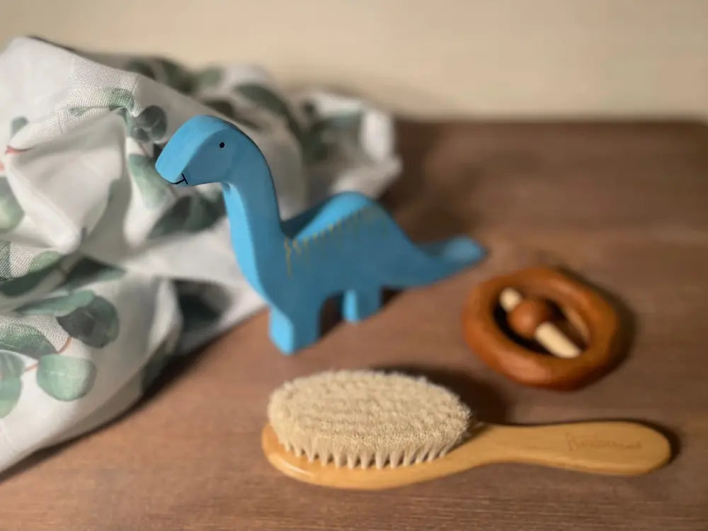 Blue dinosaur-shaped baby toy, soft bristle baby brush, and wooden toy on a wooden surface.