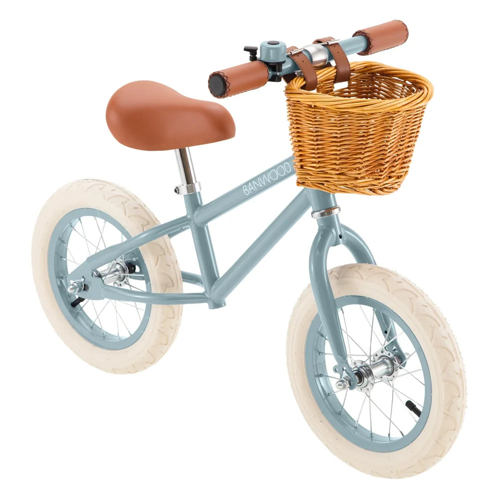 A children's balance bike with a dusty teal frame, cream tires, a brown saddle, and a natural wicker basket