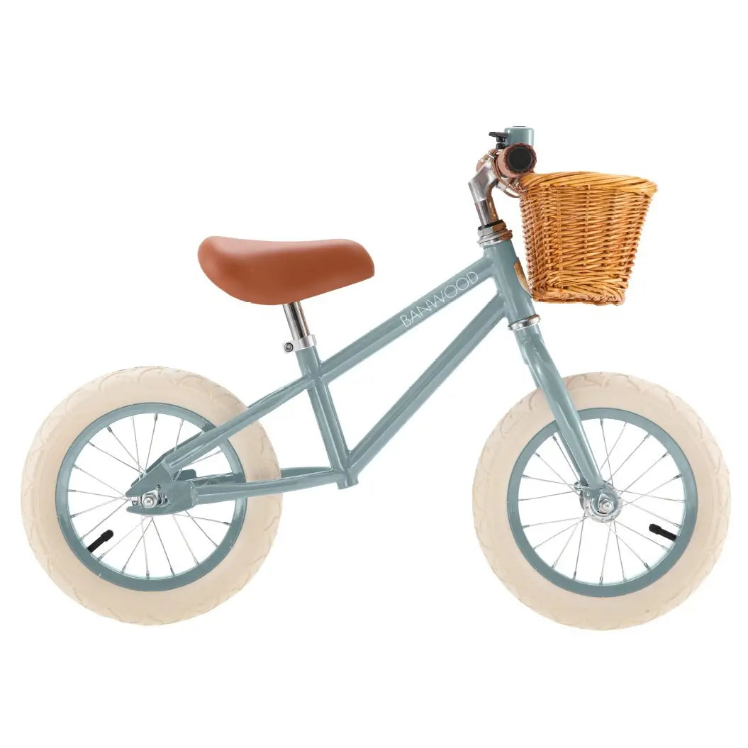 A children's balance bike in a dusty teal frame, featuring a brown saddle, a wicker basket on the front, and cream-colored tires.