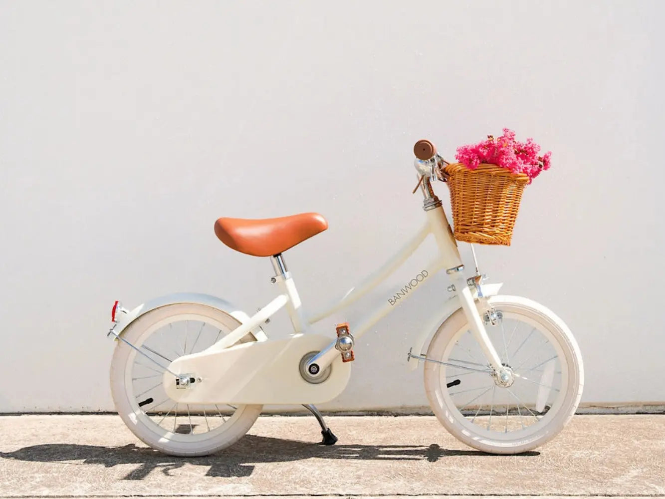 White children's bicycle with a brown seat and basket filled with pink flowers on a light gray background.