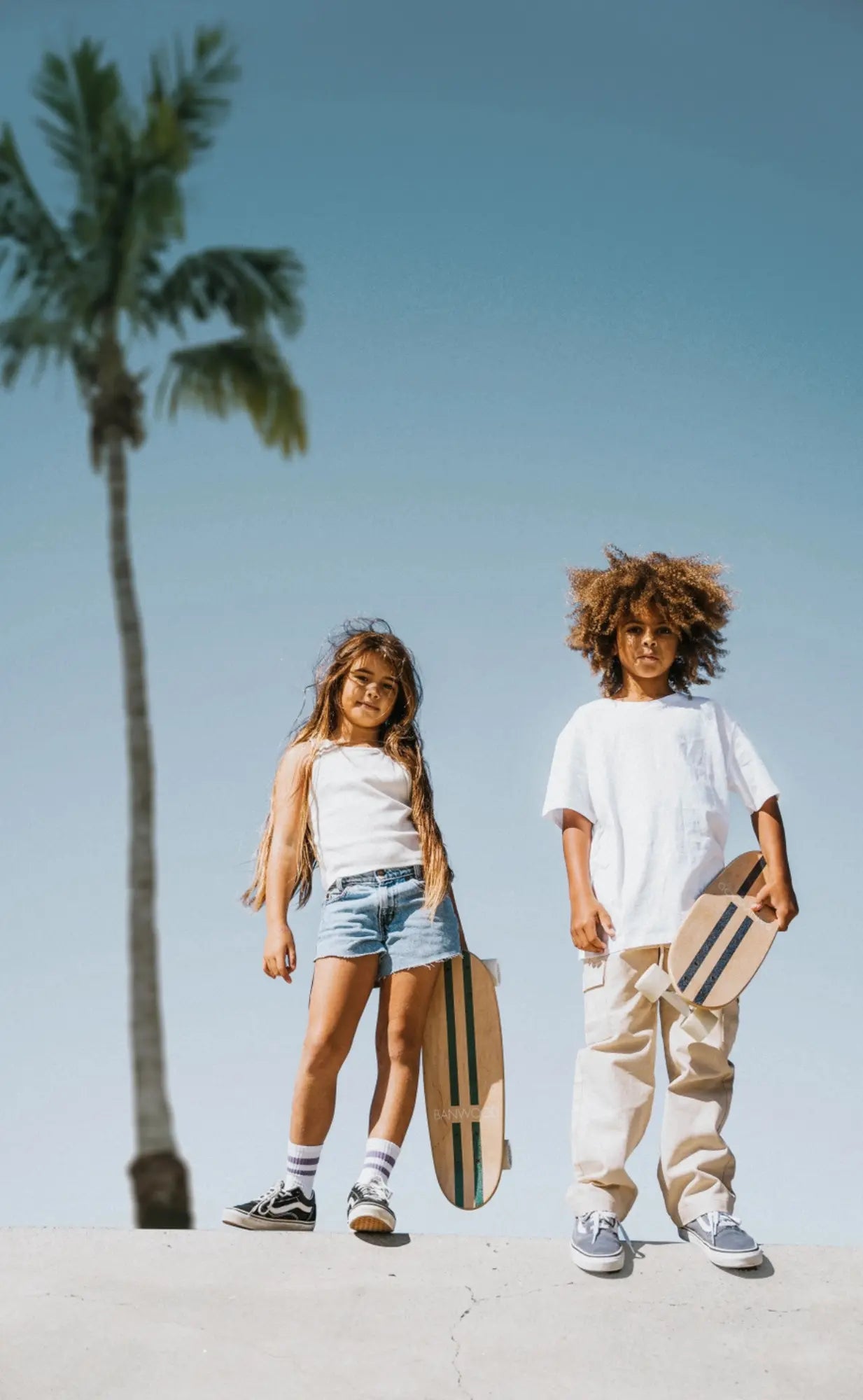 Two children with skateboards standing on a concrete surface with a palm tree in the background.