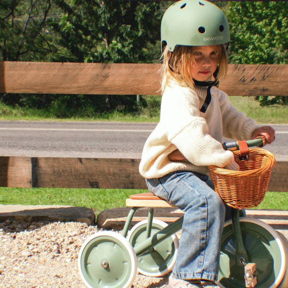 Child sitting on a tricycle with a basket, wearing a helmet, outdoors.