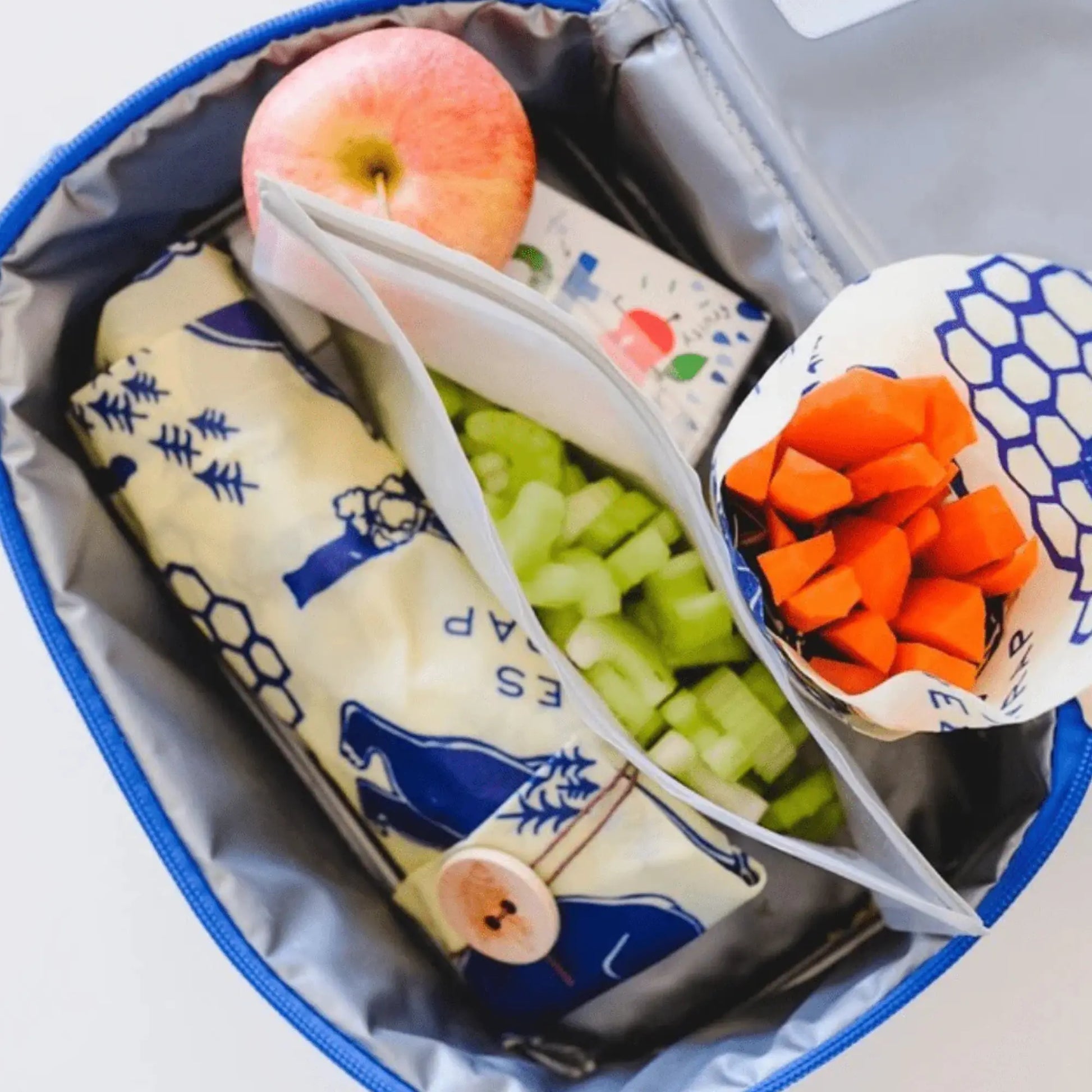 Lunchbox with cheese, apple, celery, and carrot sticks in reusable food wraps on a white background