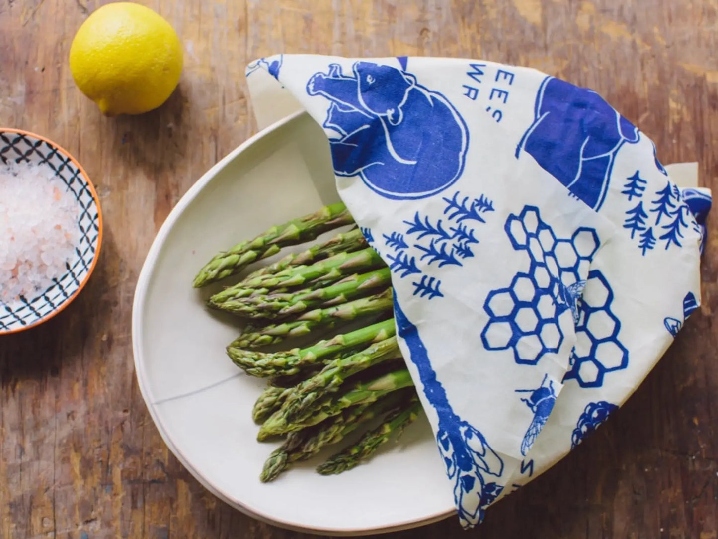 Asparagus on a plate with a blue and white patterned food wrap lemon, and salt dish on a wooden surface.
