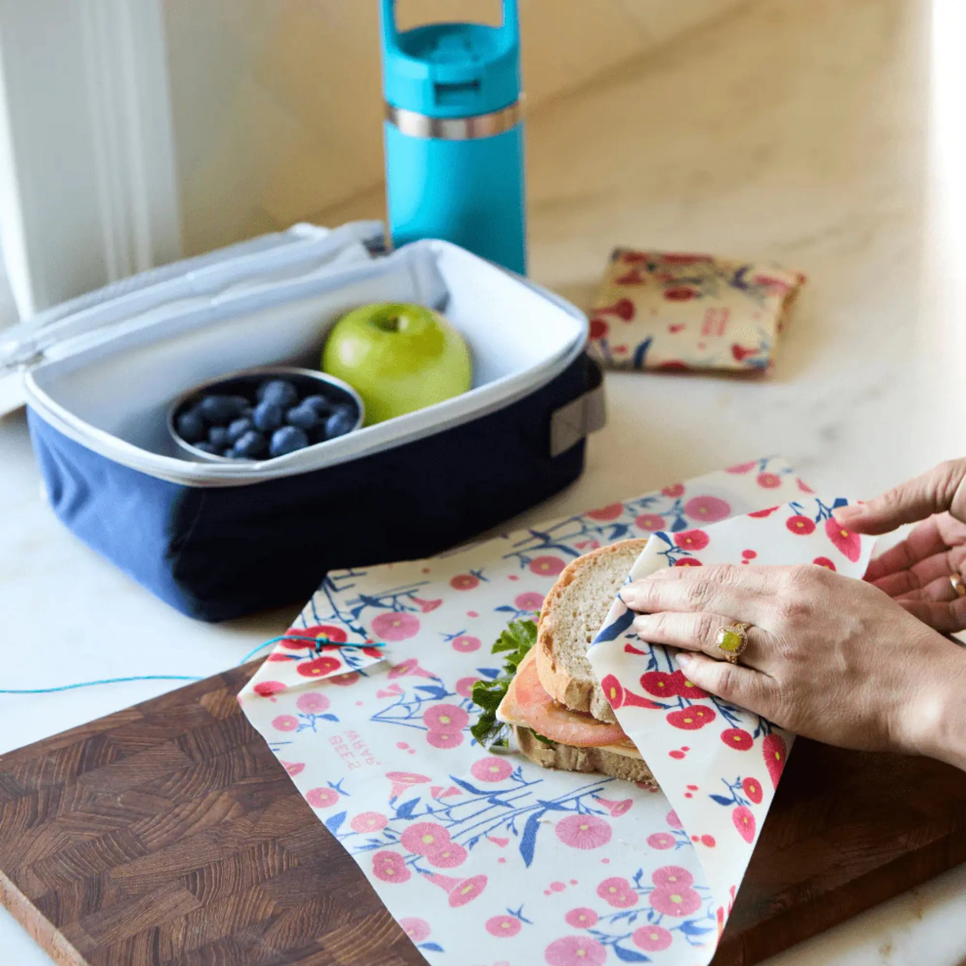 Person preparing a sandwich on floral beeswax wrap next to a lunch box with fruit and a blue thermos.