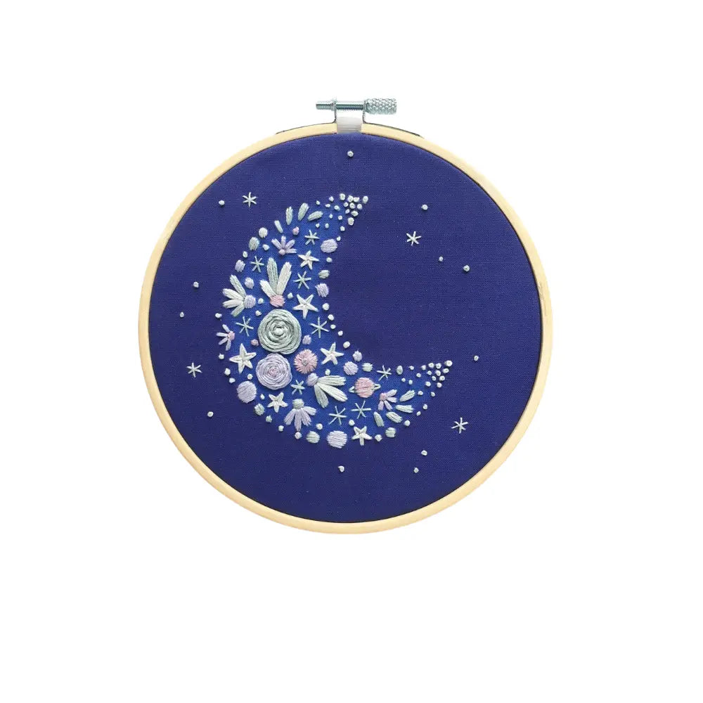Embroidery hoop with a floral moon design on a blue background, surrounded by decorative stars and marbles.