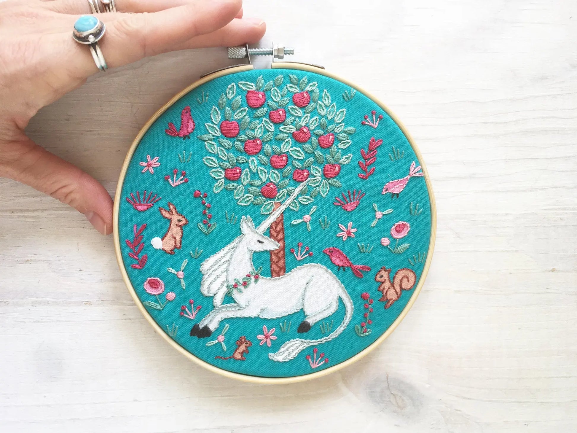Embroidery hoop with a unicorn and floral, woodland animal, apple tree design on a teal background