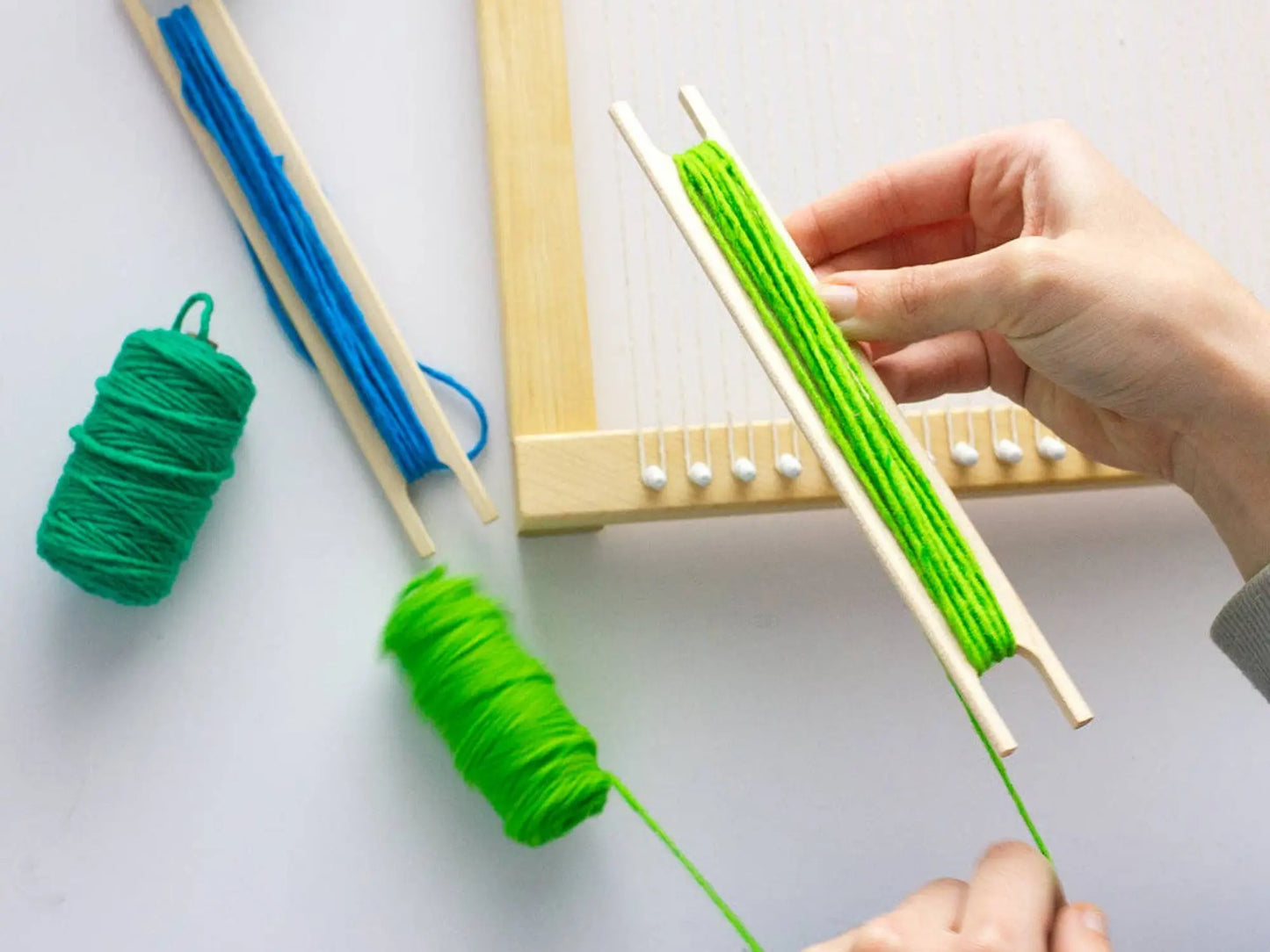 Person using a small wooden loom with green yarn on a white background