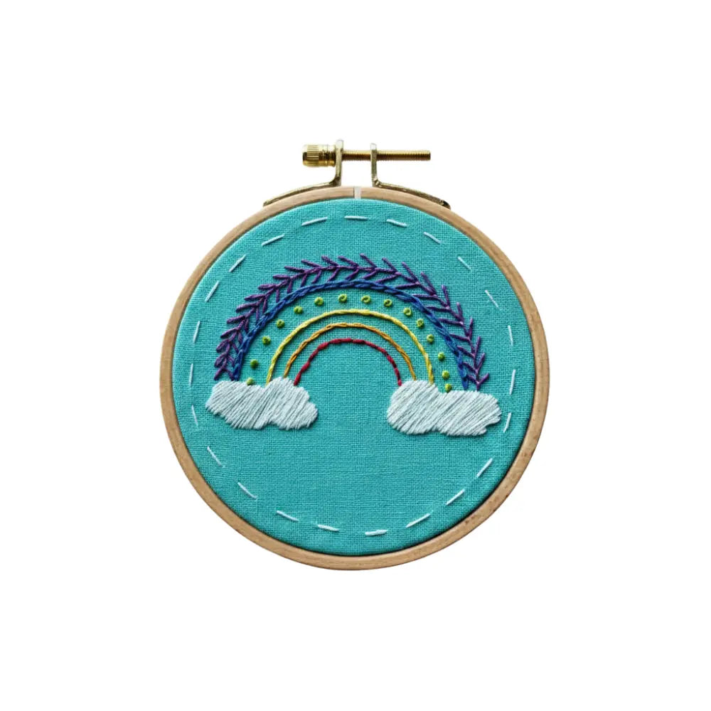 Embroidery hoop with a rainbow and cloud design on teal fabric.