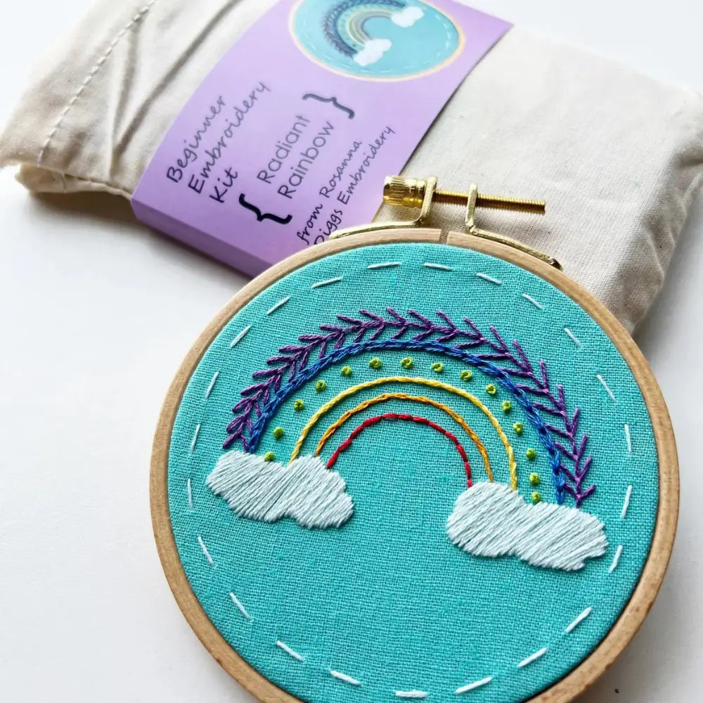 Embroidery hoop with rainbow design on teal fabric, surrounded by a kit packaging.