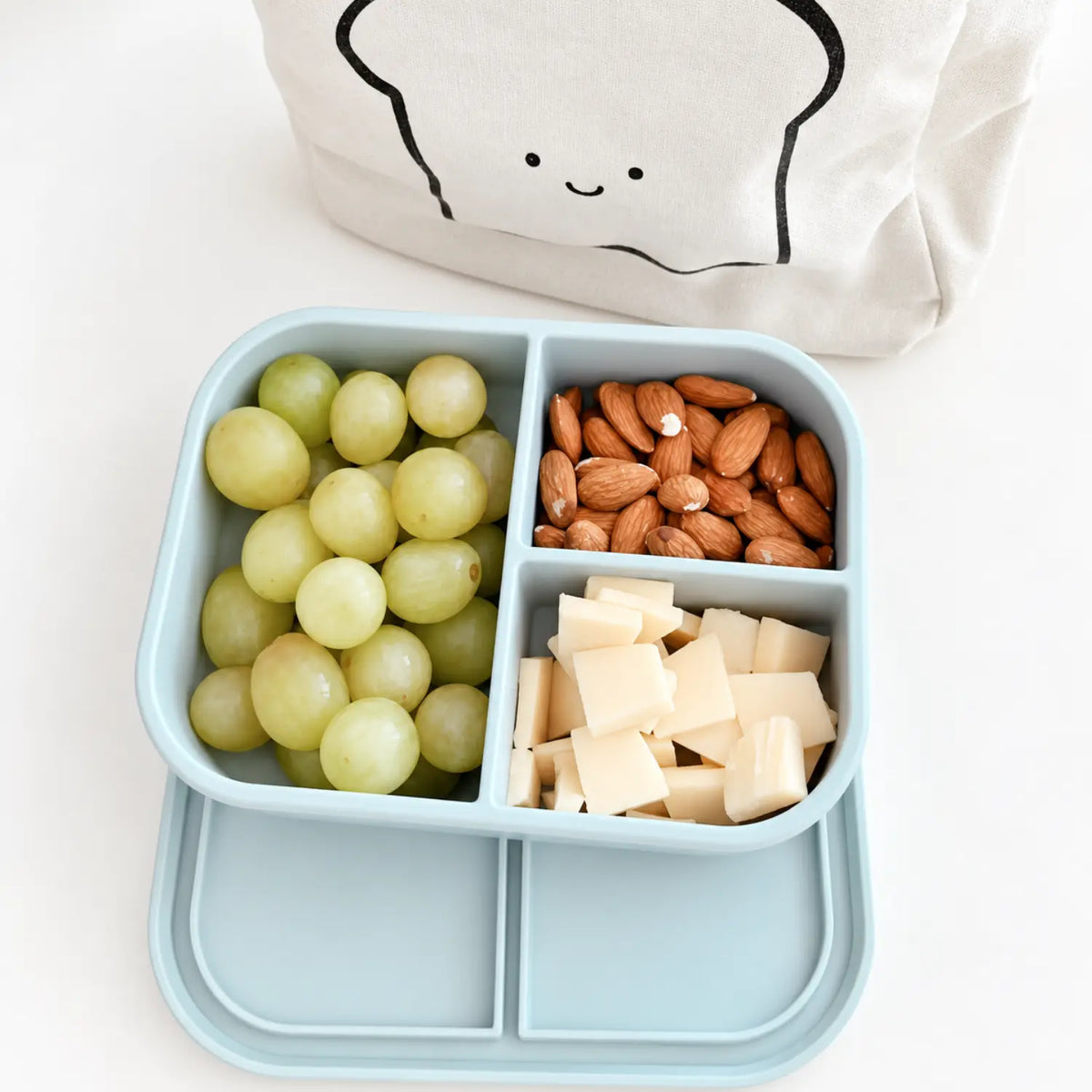 Light blue Bear silicone bento box packed with snacks and lunch foods beside a lunch bag.
