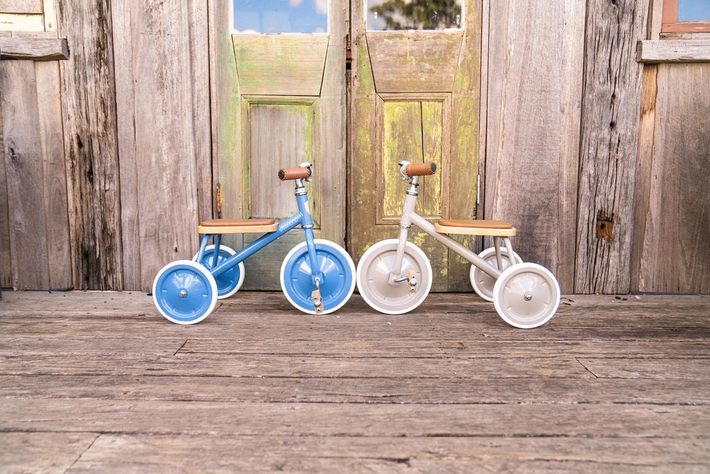 Two children's tricycles, one blue and one white, on a wooden deck.