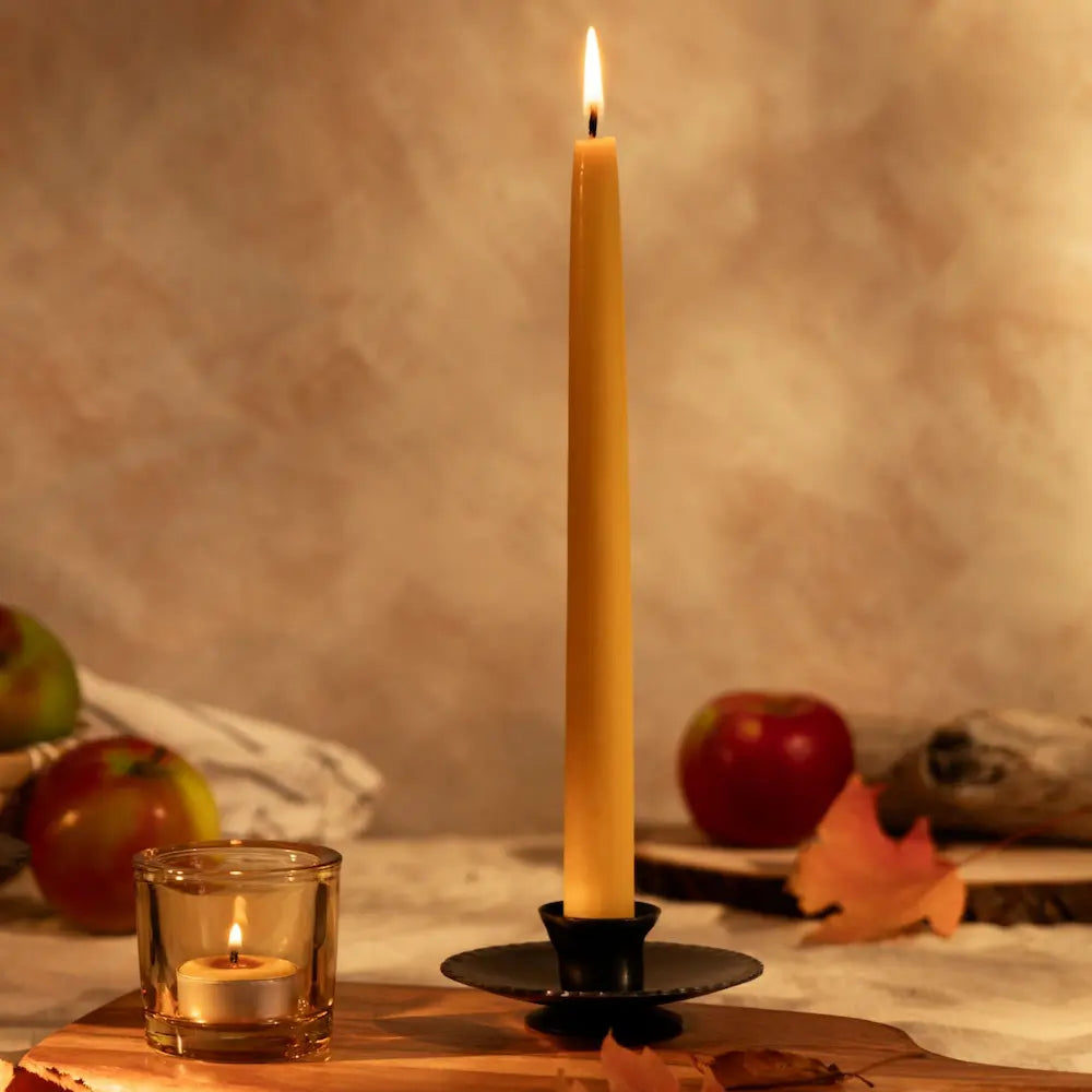 Tall lit candle on a wooden board with apples and a small candle in the background.