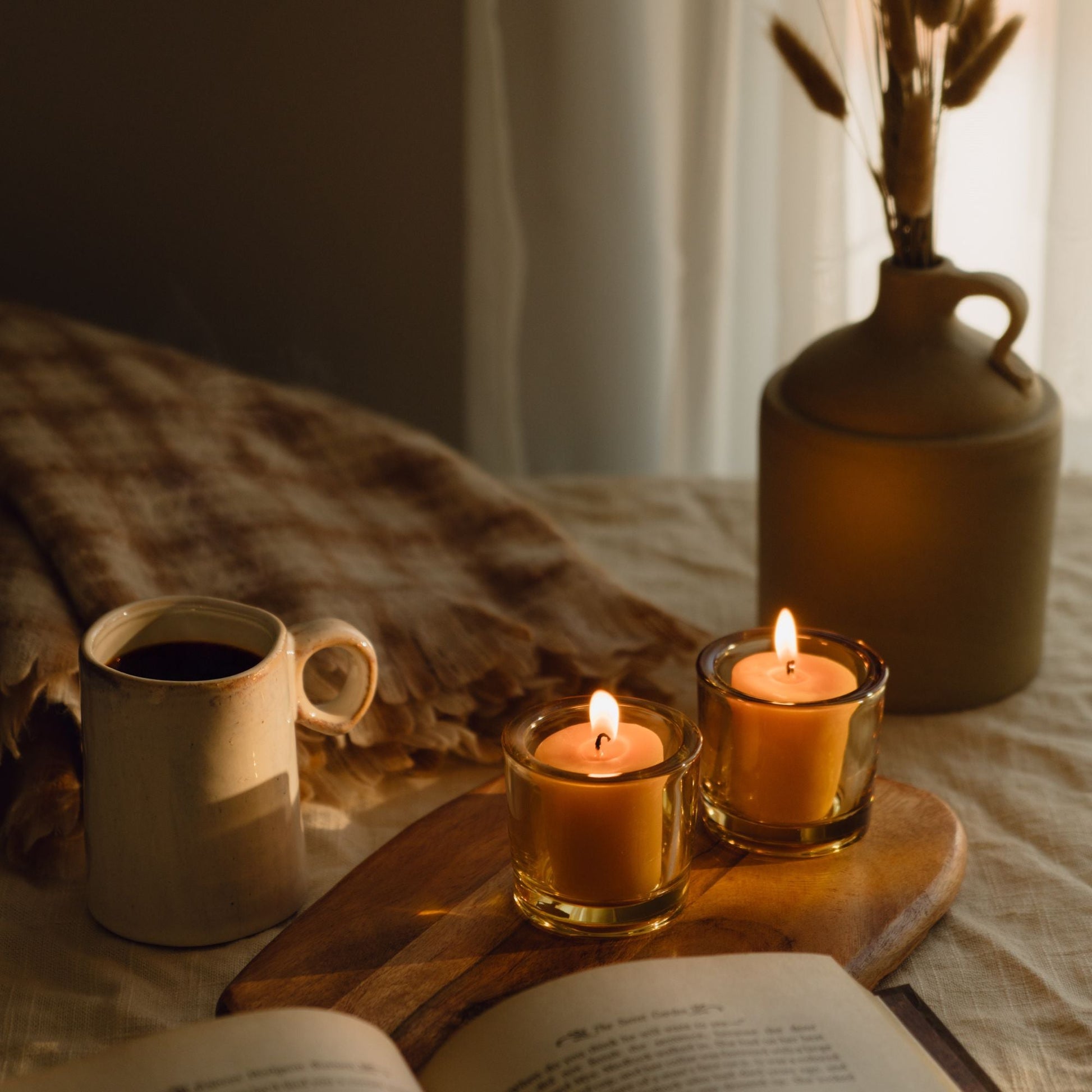 Candlelit scene with a mug, book, and vase on a wooden board.
