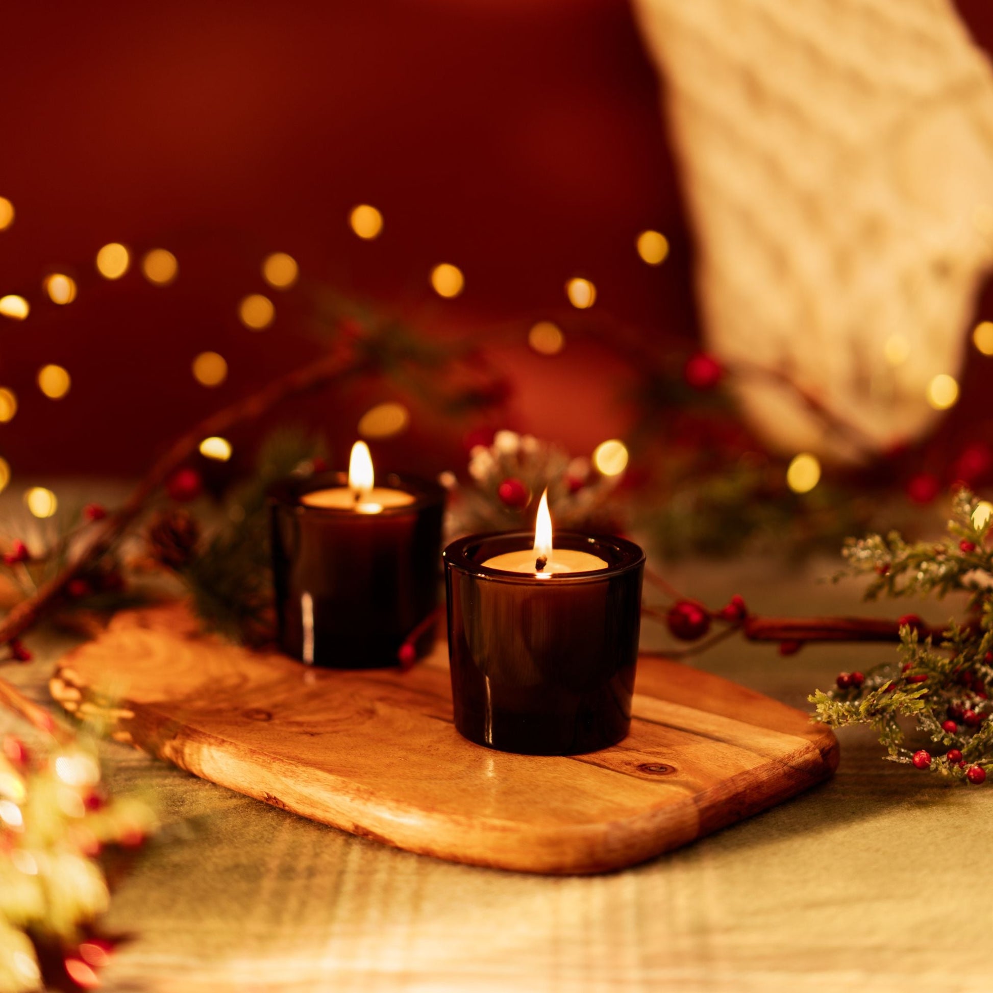 Two lit candles on a wooden board with blurred lights and decorations in the background