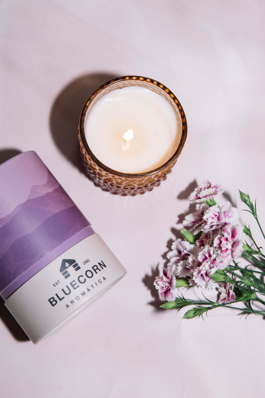 Candle in a glass holder with a purple container labeled 'Bluecorn Aromatica' on a light pink background.