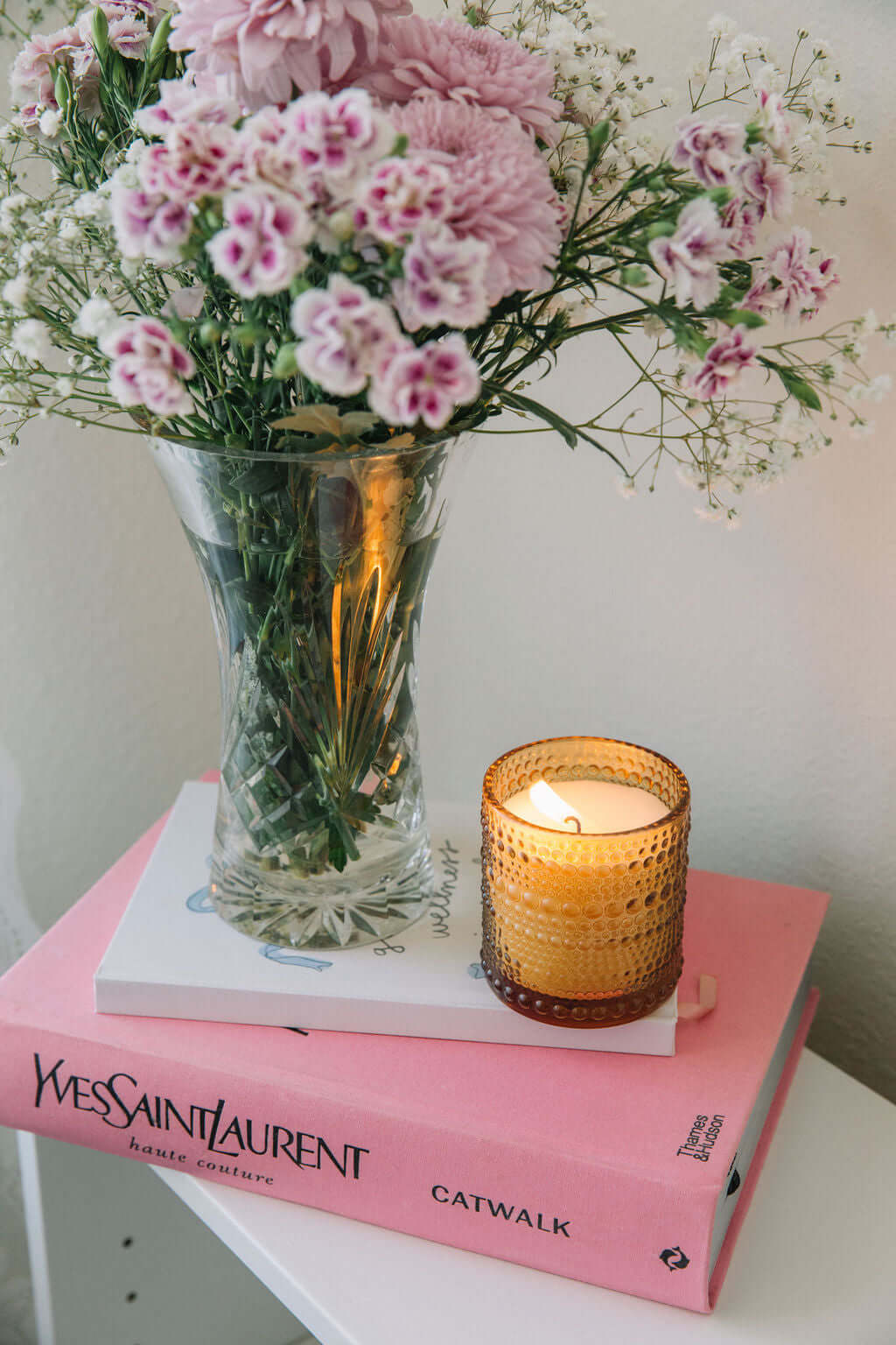 Vase with pink flowers and a lit candle on a pink book titled 'Catwalk' by Yves Saint Laurent.
