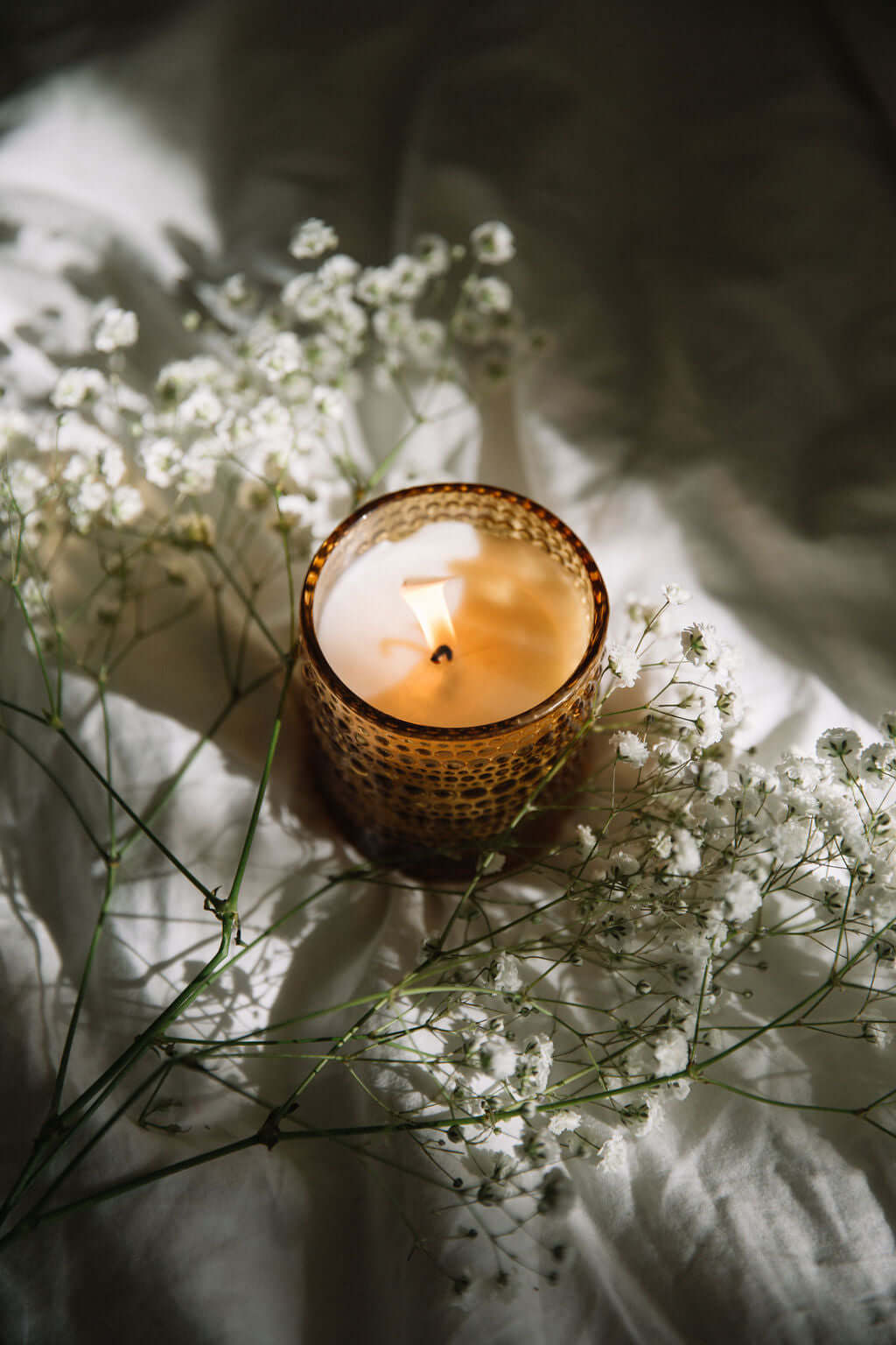 Candle in a textured holder surrounded by delicate white flowers on a soft surface