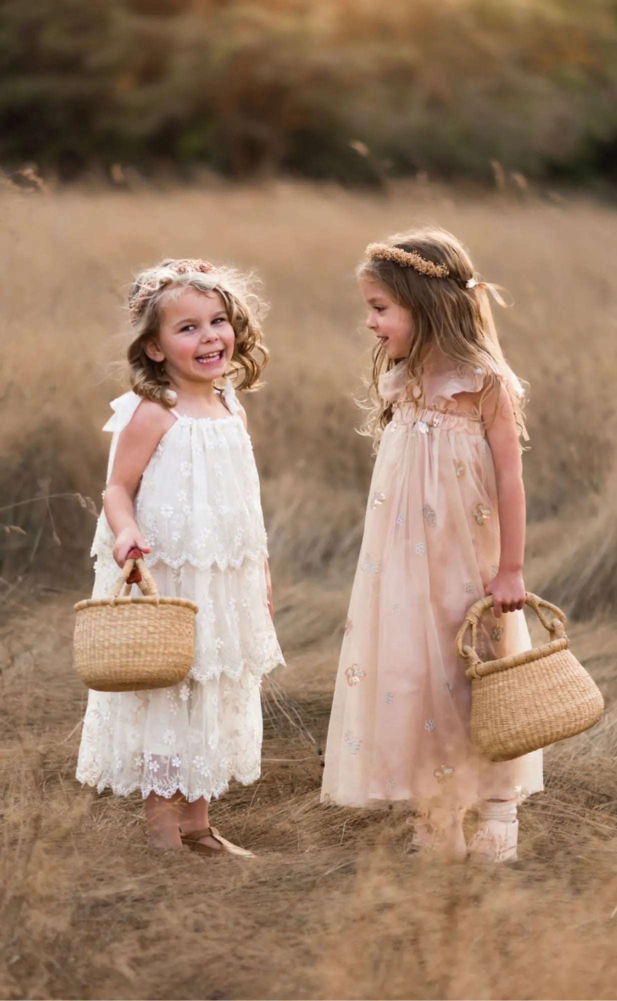 Two young girls in floral dresses standing in a field holding wicker bags.