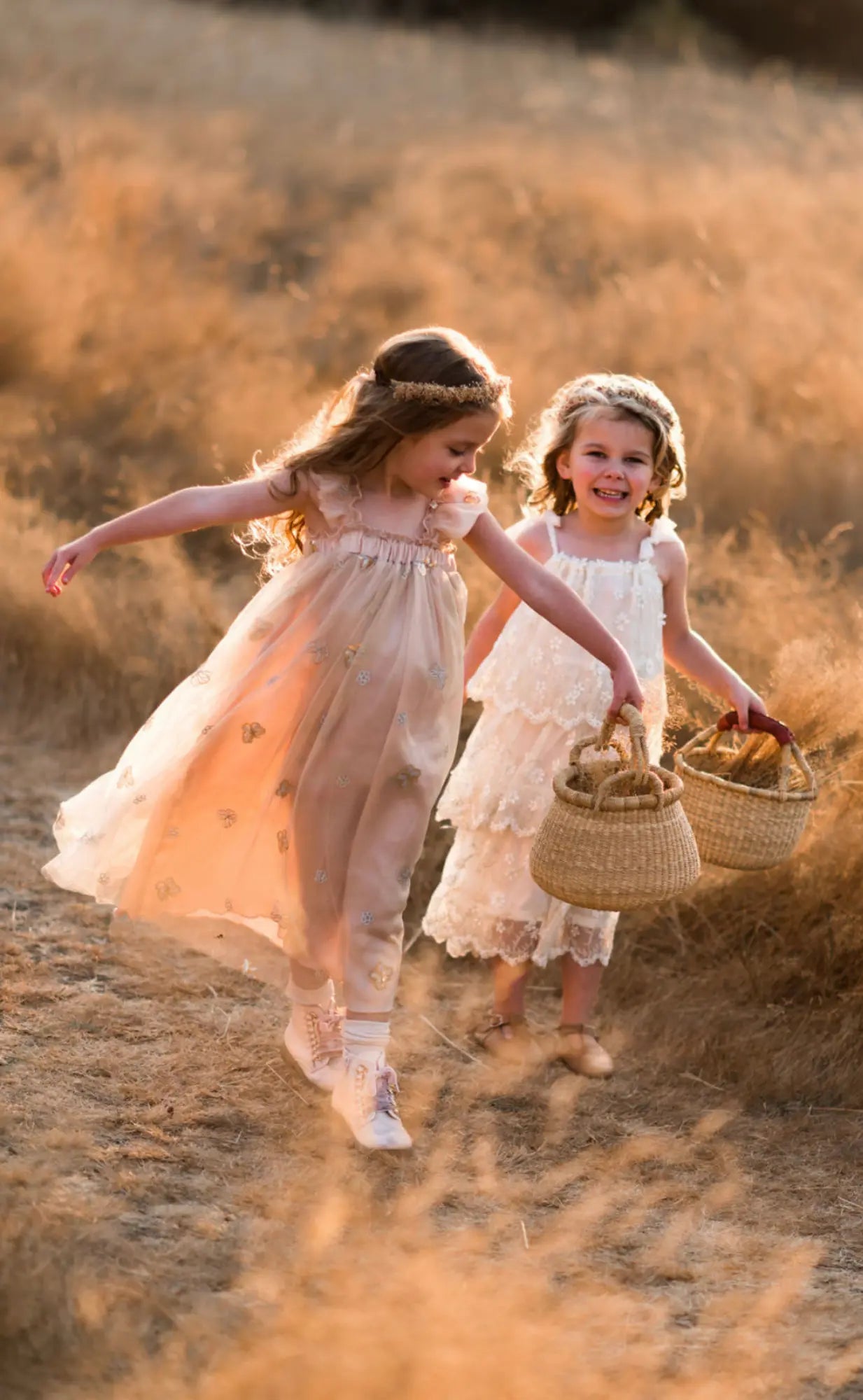 Two young girls in dresses walking through a field with baskets.