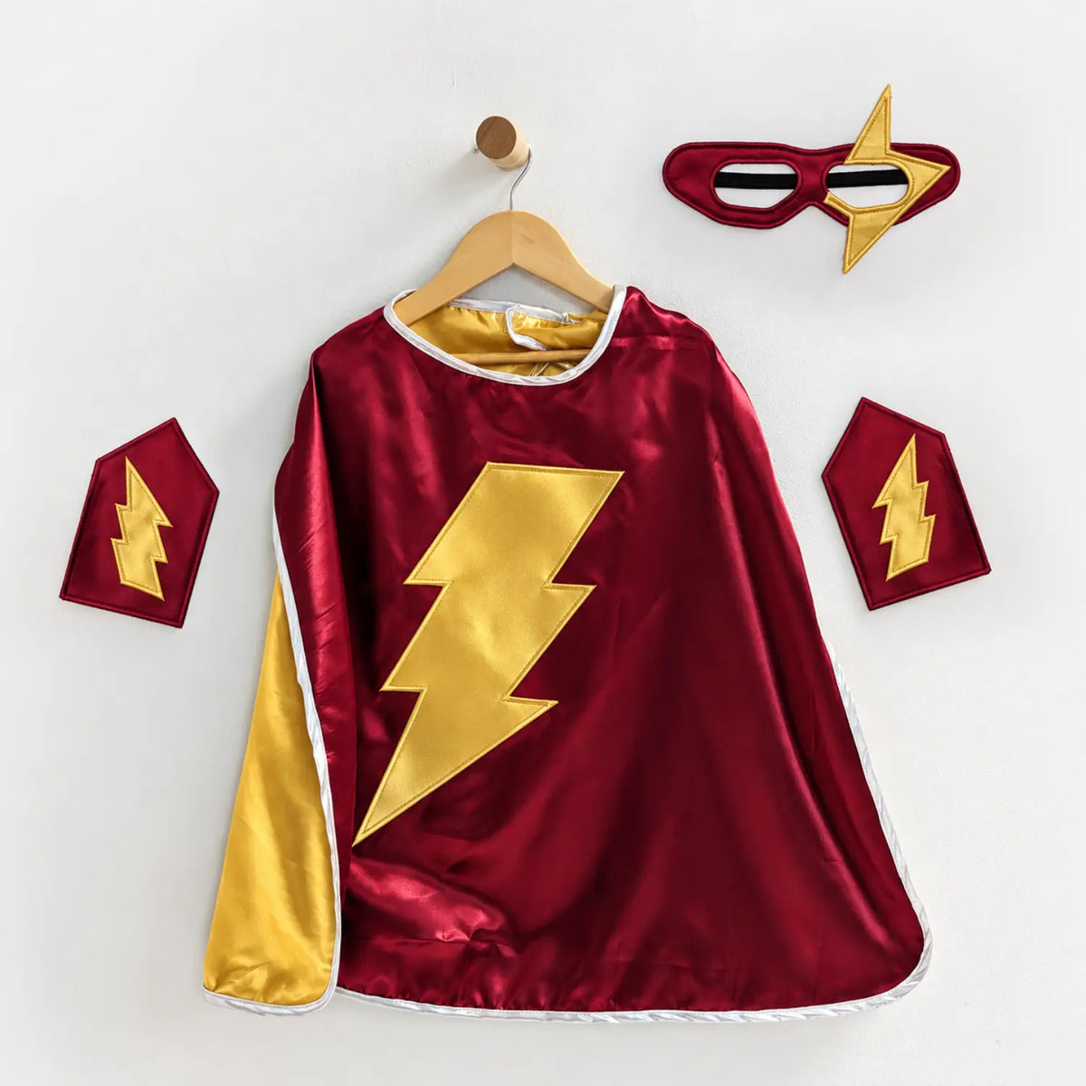 Burgundy superhero cape set with gold lightning bolt emblem, matching eye mask, and wrist cuffs.