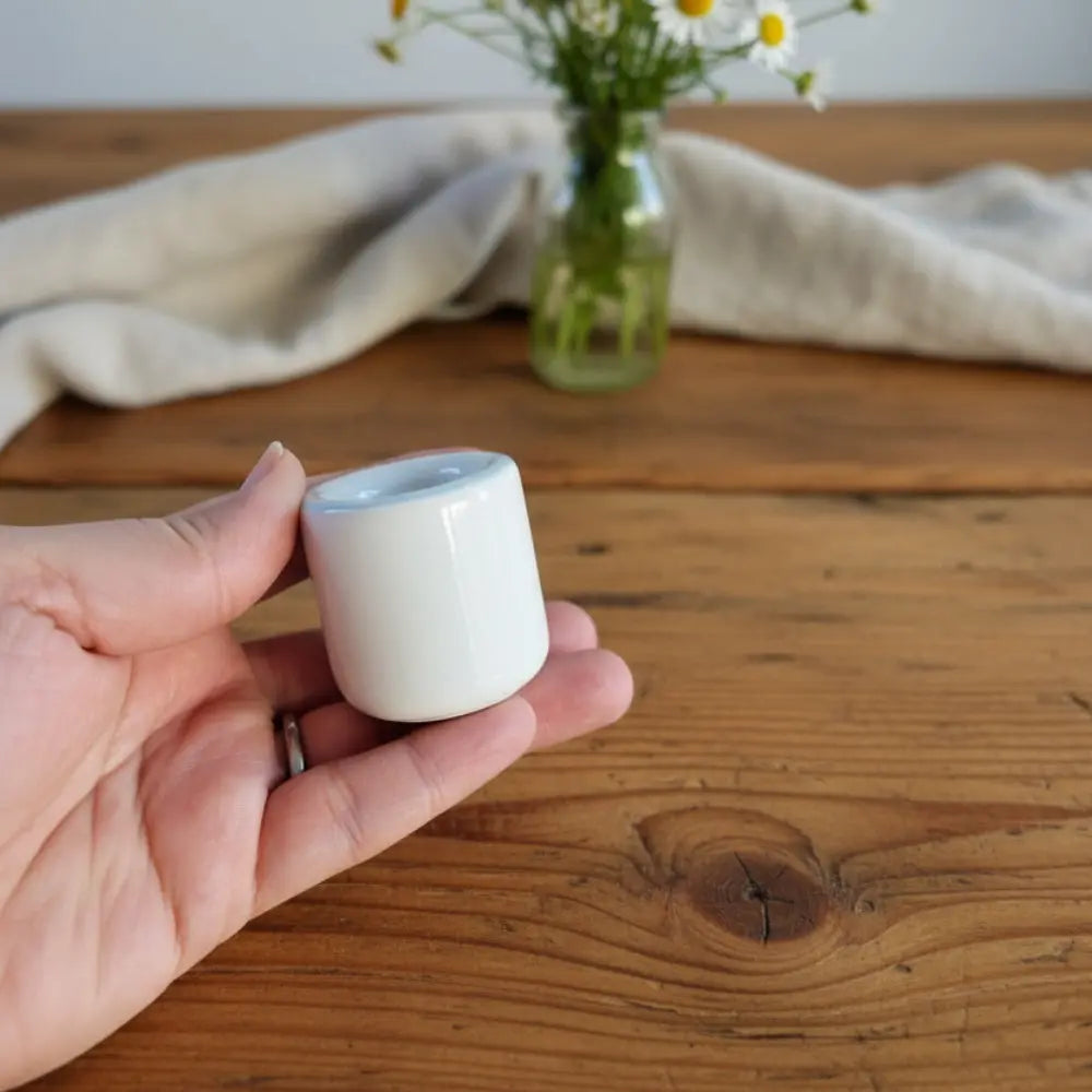 Hand holding a small white ceramic taper candle holder on a wooden surface with a vase of flowers 