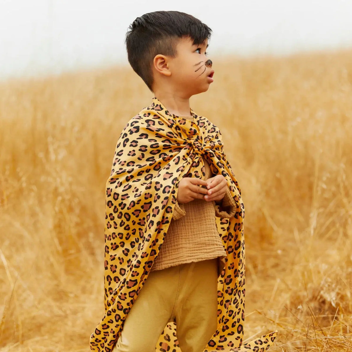 Child in a cheetah playsilk cape looking out across tall dry grass during outdoor pretend play