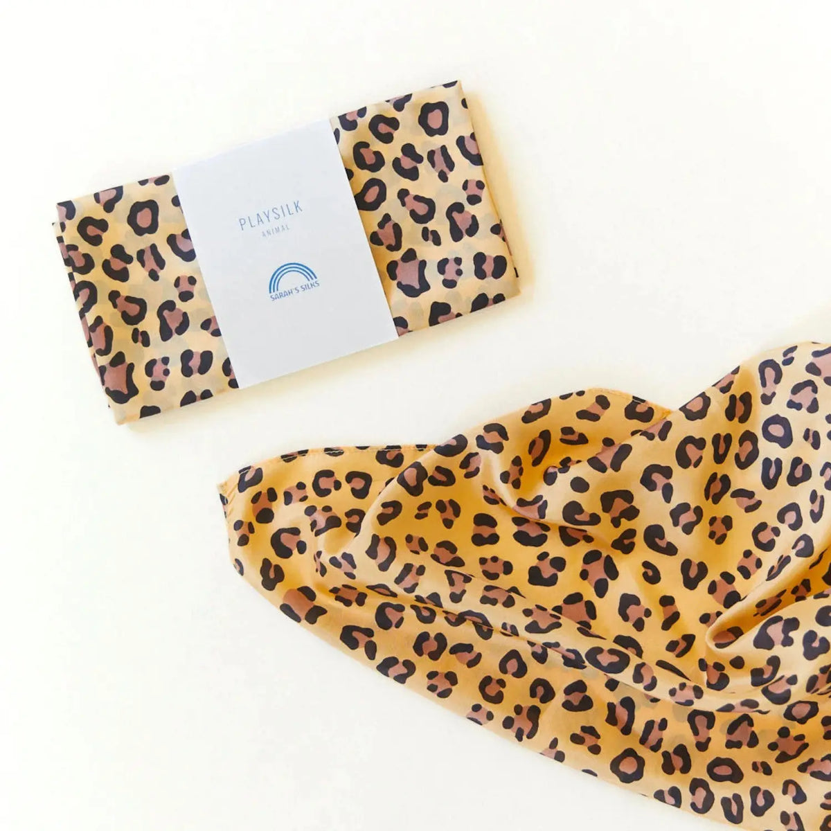 Cheetah print playsilk folded in its Sarah’s Silks paper band beside an unfolded silk