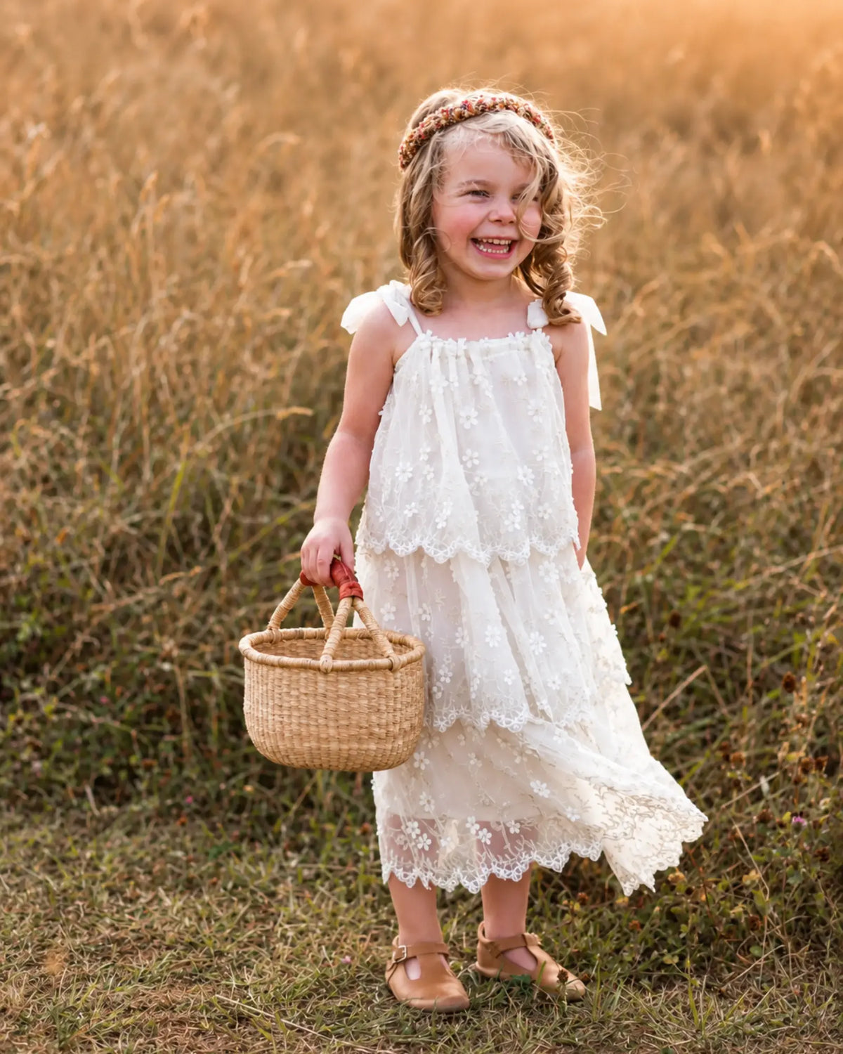 Young girl in a white embroidered dress smiling as she holds out a small handwoven Bolga gathering basket in a sunlit field.