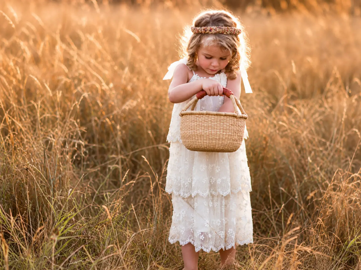 Young girl wearing a flower crown looking inside her handwoven Bolga basket while standing among golden meadow grasses.
