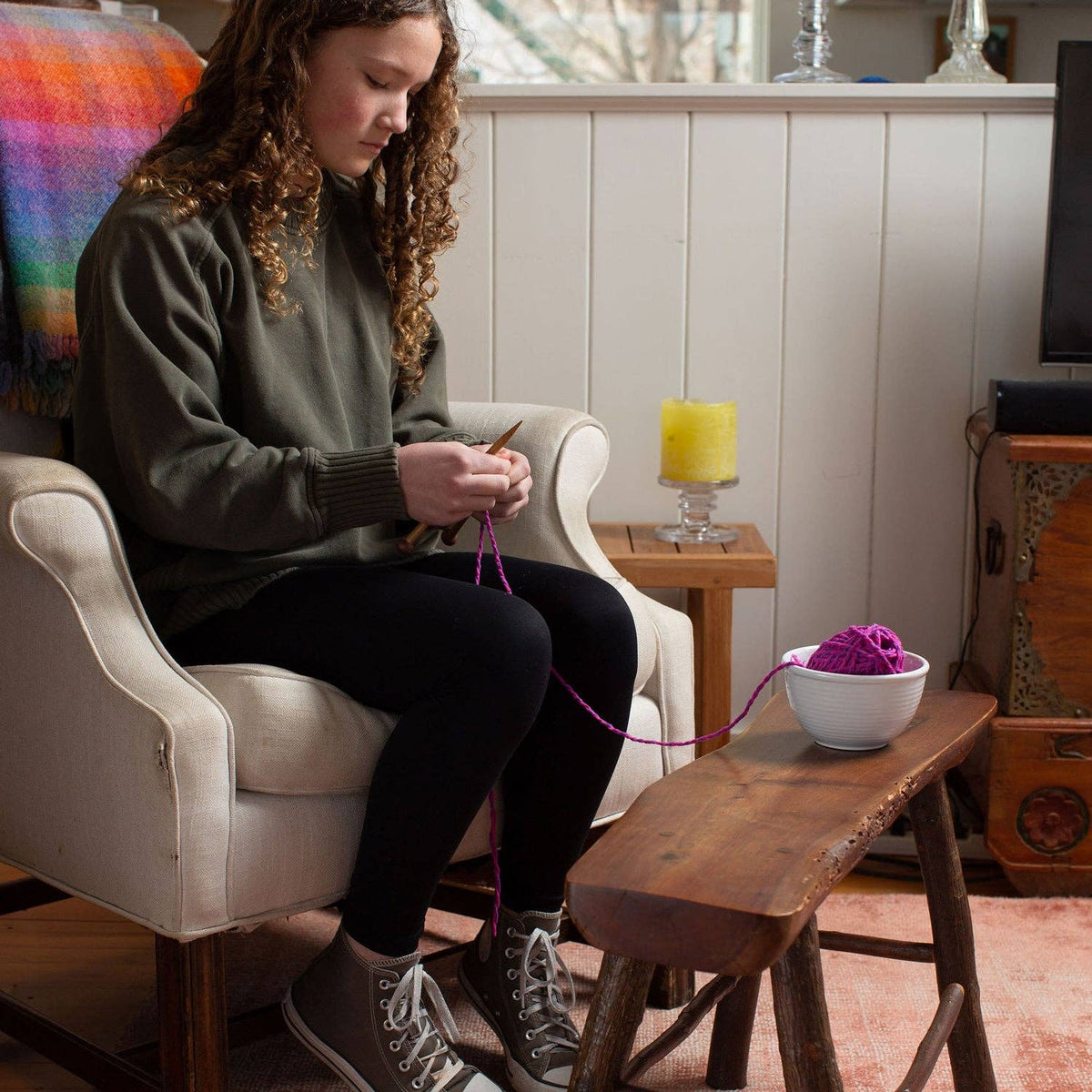 Young knitter sitting in an armchair working on a wool scarf with wooden needles and a ball of yarn in a bowl