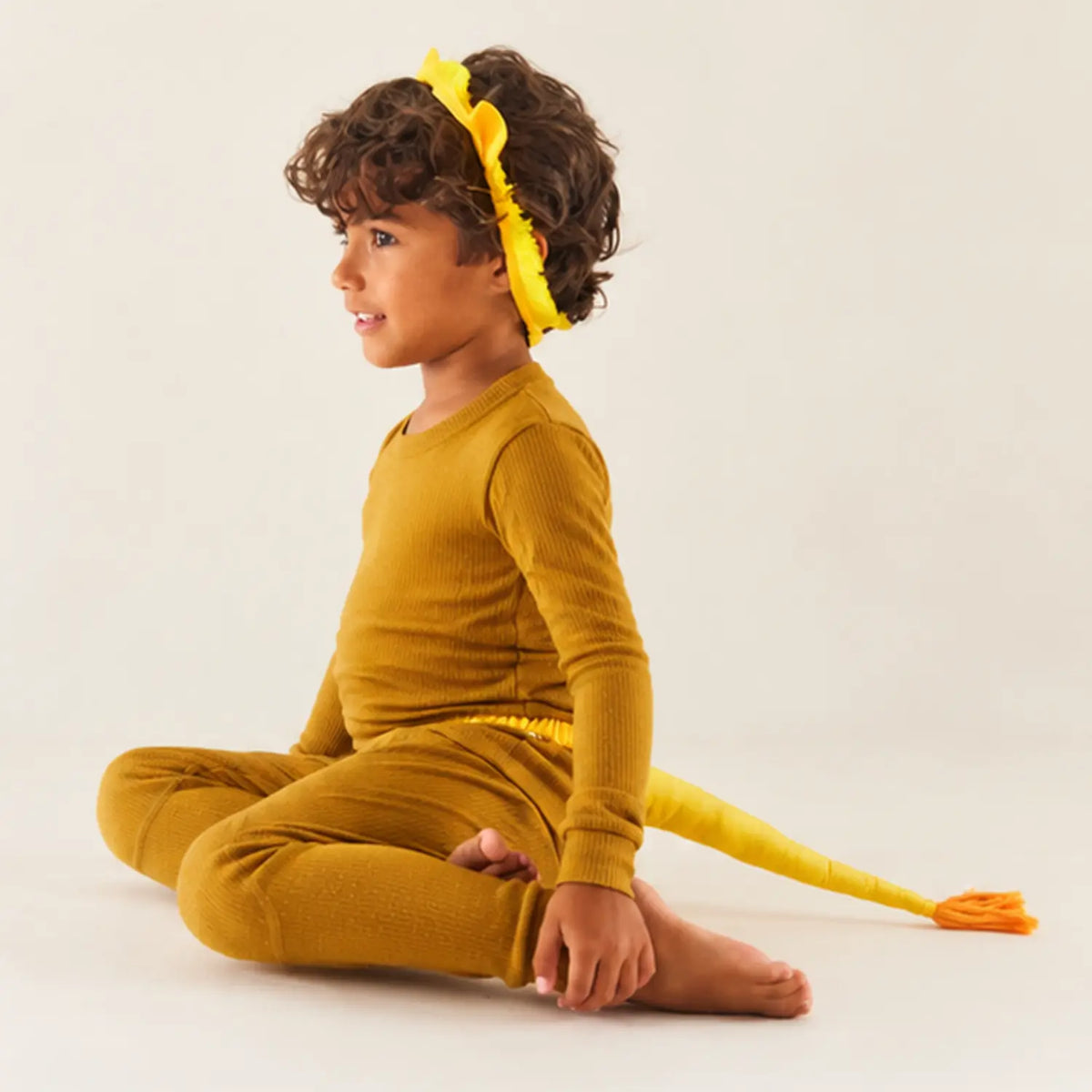 Child sitting on the floor in a lion costume with lion ears and a yellow lion tail with an orange tassel