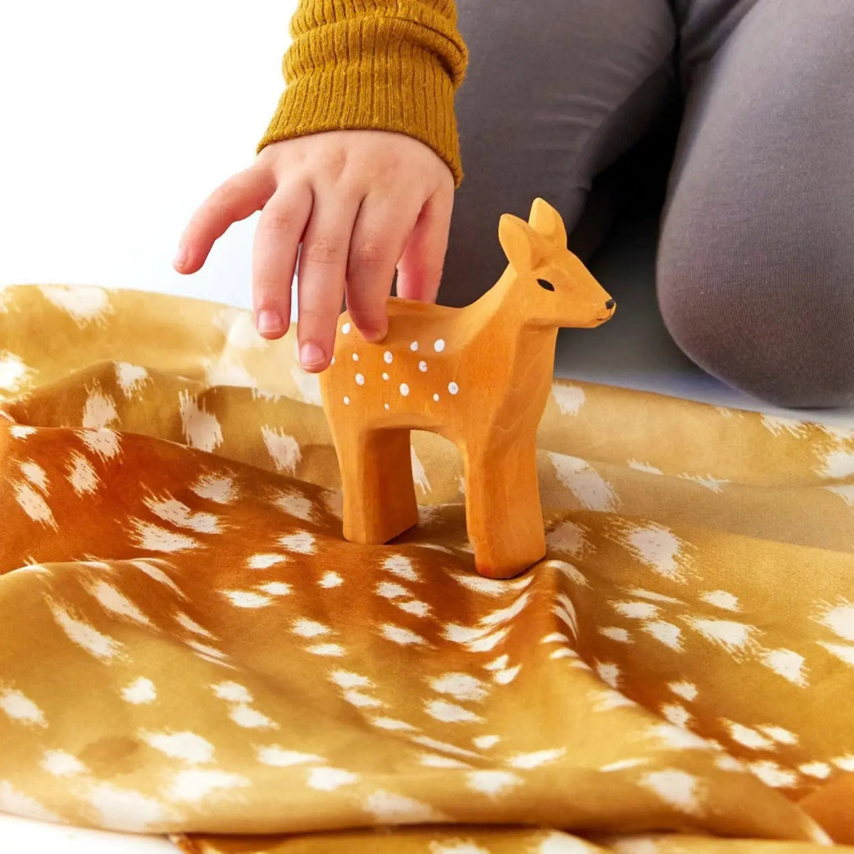 A child’s hand guides a wooden fawn across a golden fawn-print Playsilk