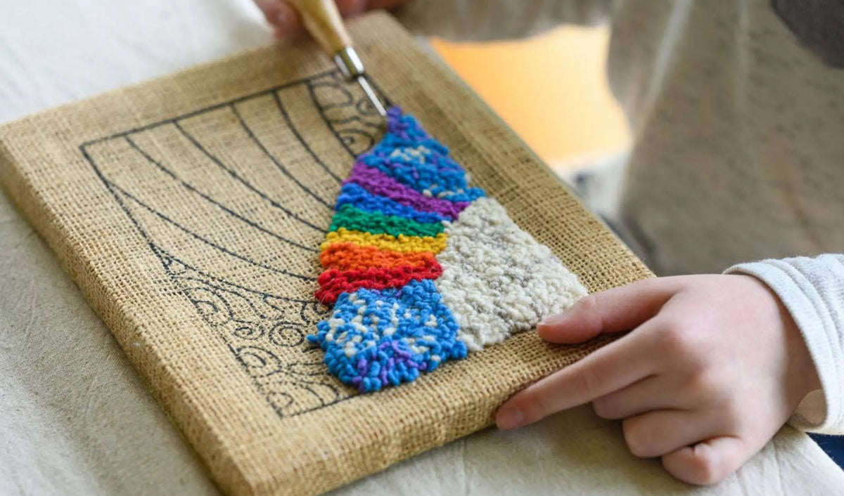 Child using a rug hook to pull loops of rainbow wool yarn through burlap on a wooden frame