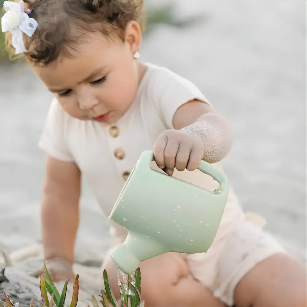 Child holding a green silicone watering can pouring water on plants in the sand