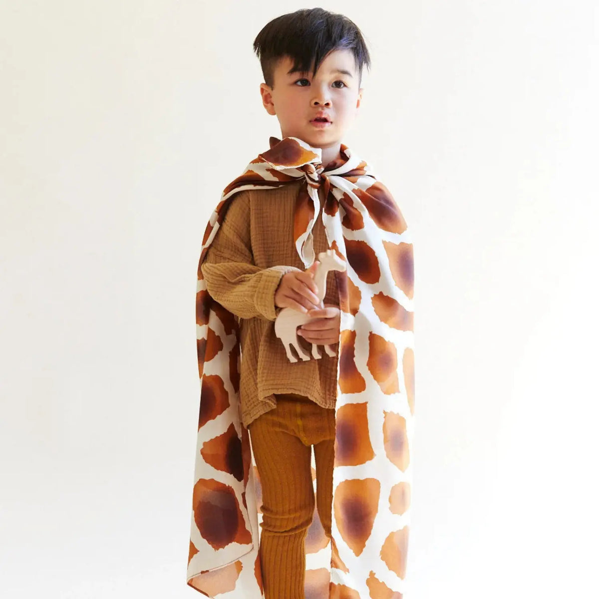 Child wearing a giraffe print Playsilk as a cape and holding a wooden giraffe toy for pretend play