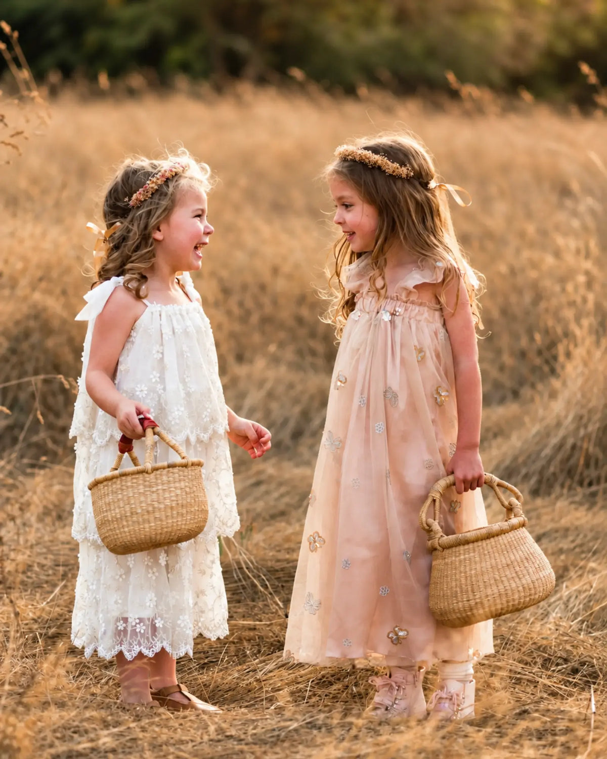 Two girls wearing flower crowns and carrying natural woven Bolga baskets while sharing a laugh in a grassy meadow.