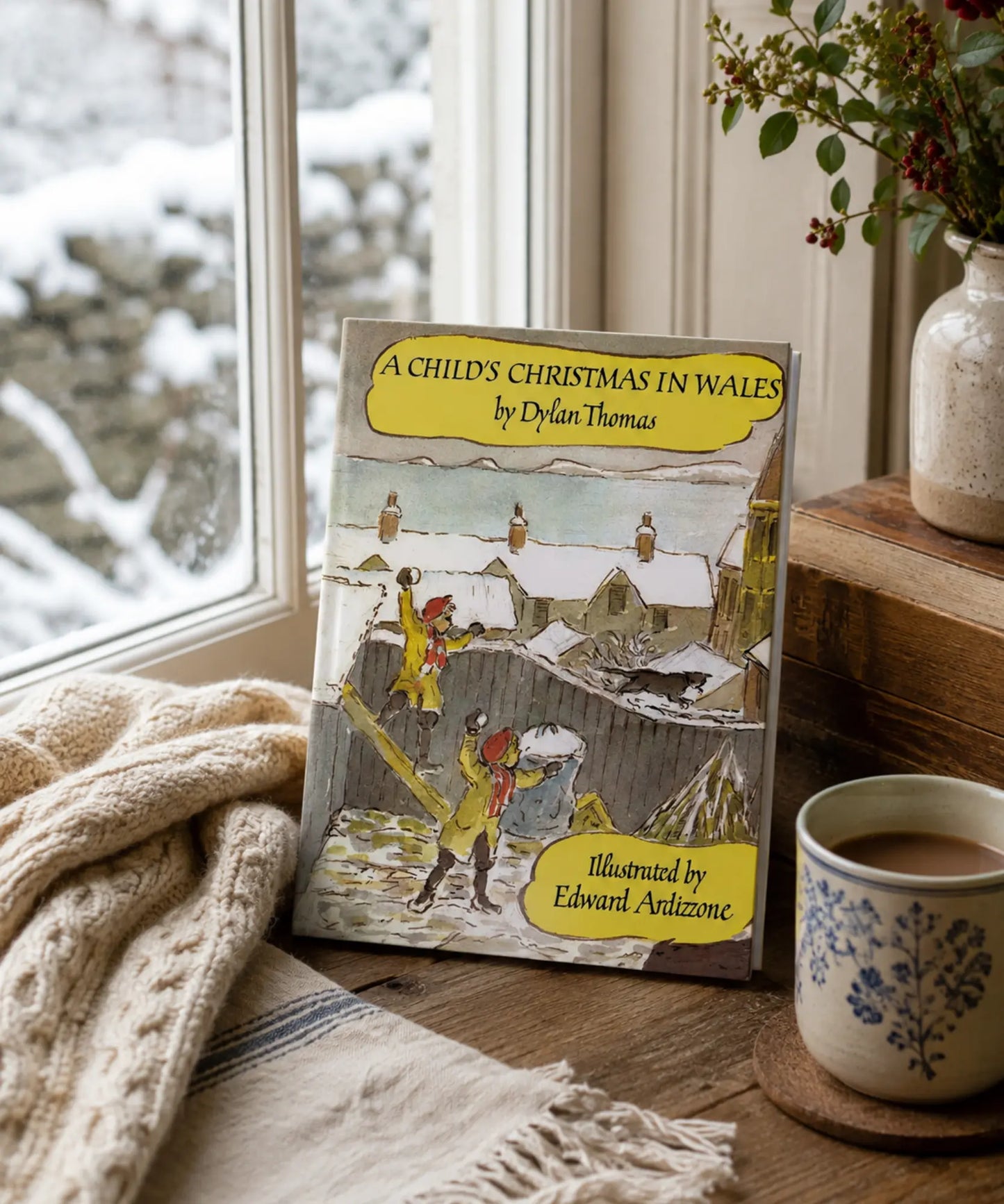 Book titled 'A Child's Christmas in Wales' by Dylan Thomas on a windowsill with a cup of coffee and blanket.