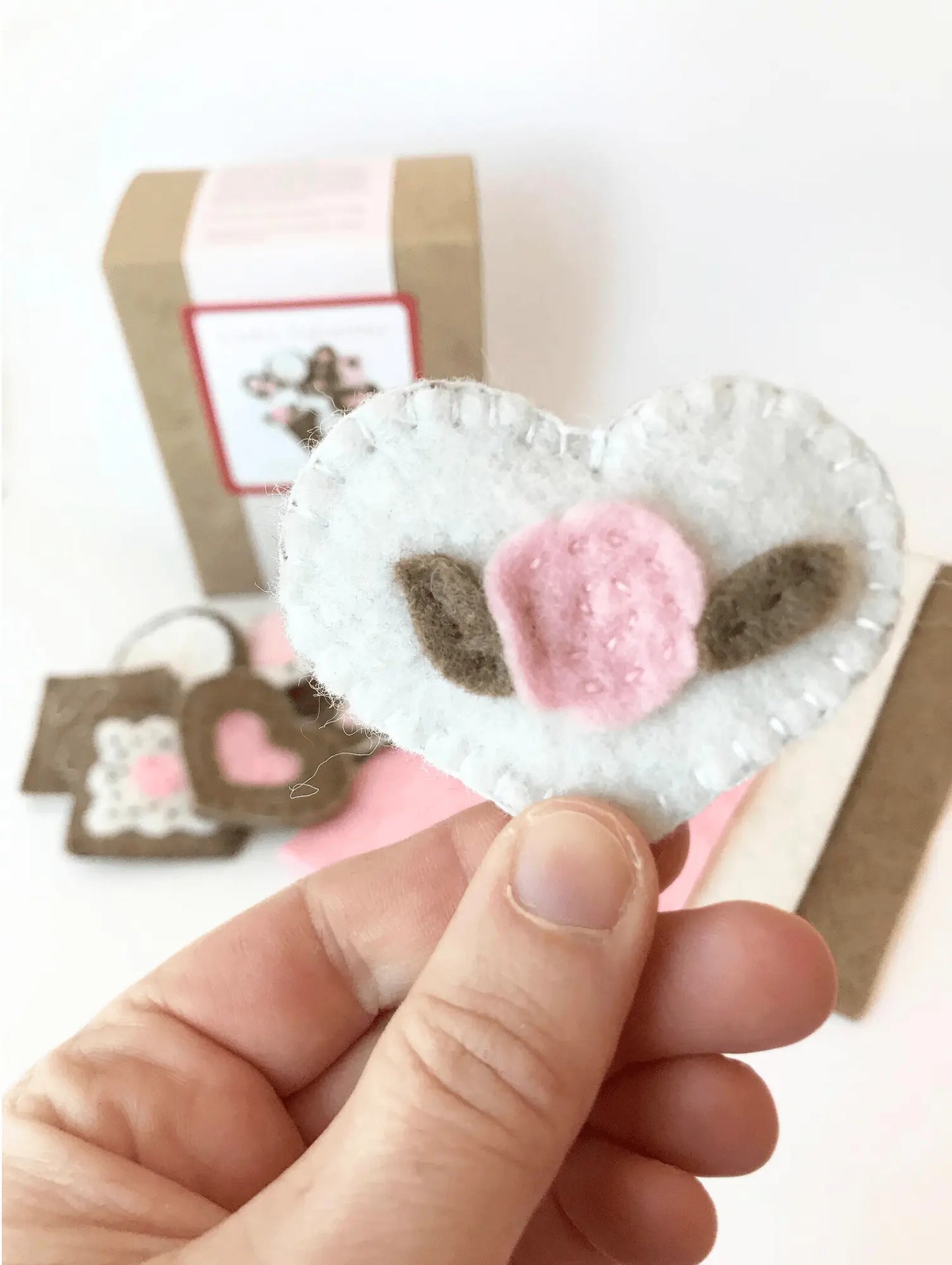 Heart-shaped felt craft with pink flower and brown leaves held by a hand on a white background