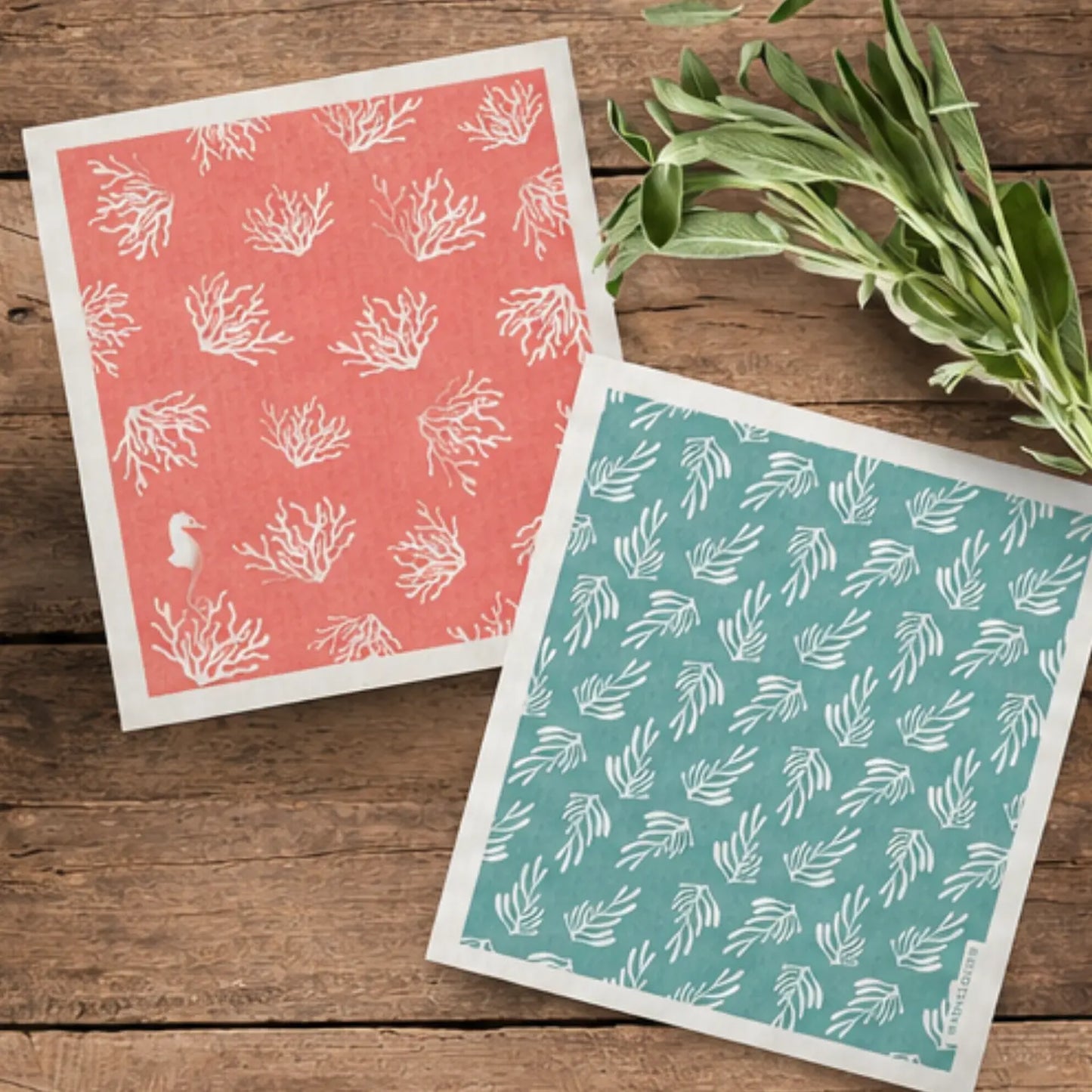 Coral and kelp Swedish dishcloth pair arranged on a rustic wood surface with fresh herbs