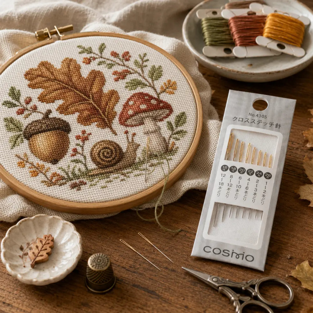 Embroidery hoop with autumn-themed design, threads, scissors, and a package of needles on a wooden surface.