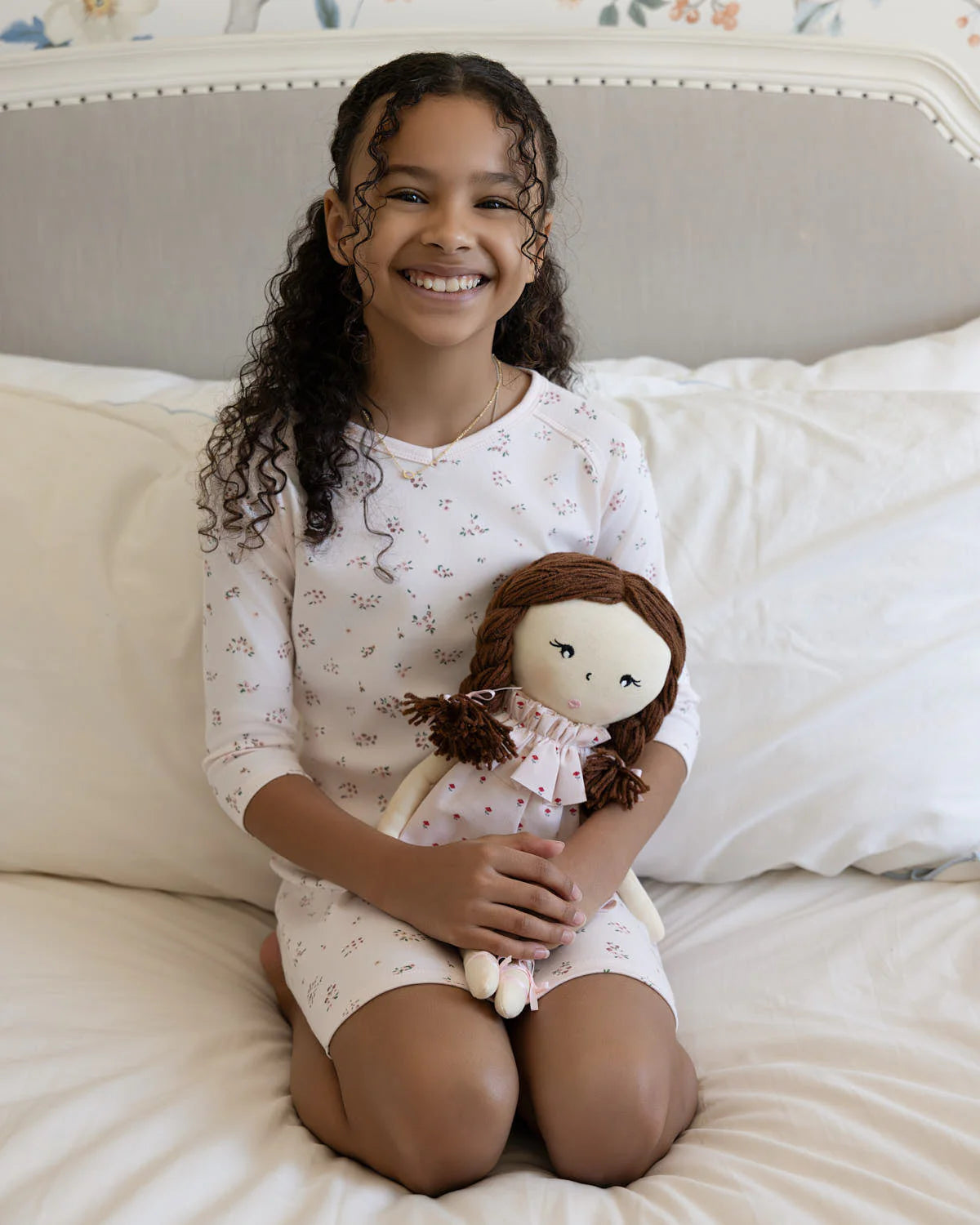 Young girl sitting on a bed holding a doll, wearing a floral dress.
