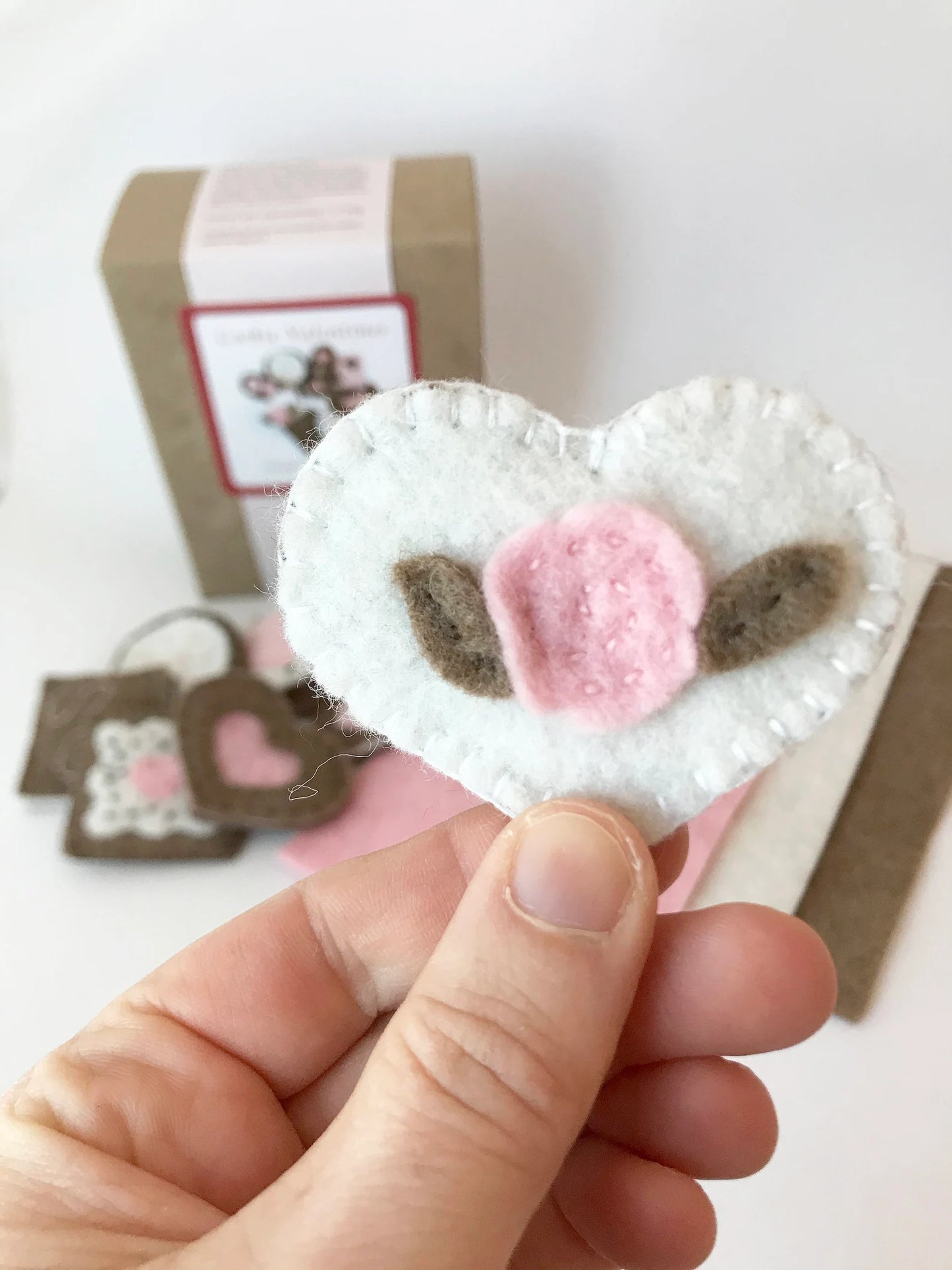 Heart-shaped felt craft with pink flower and brown leaves held by a hand on a white background